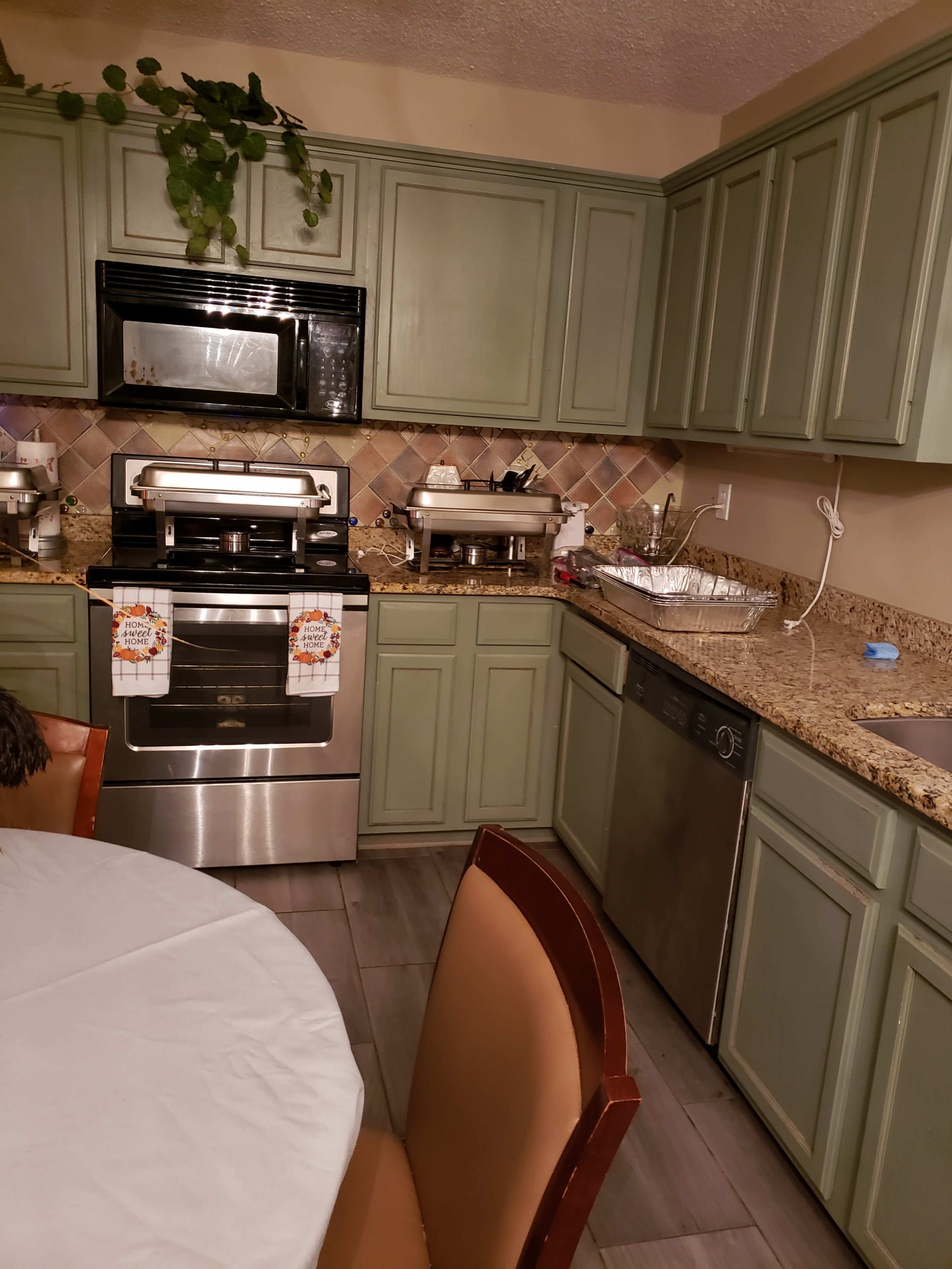 A kitchen with green cabinets, a stainless steel oven, and a round table with brown chairs, along with various appliances and kitchen items on the countertops.