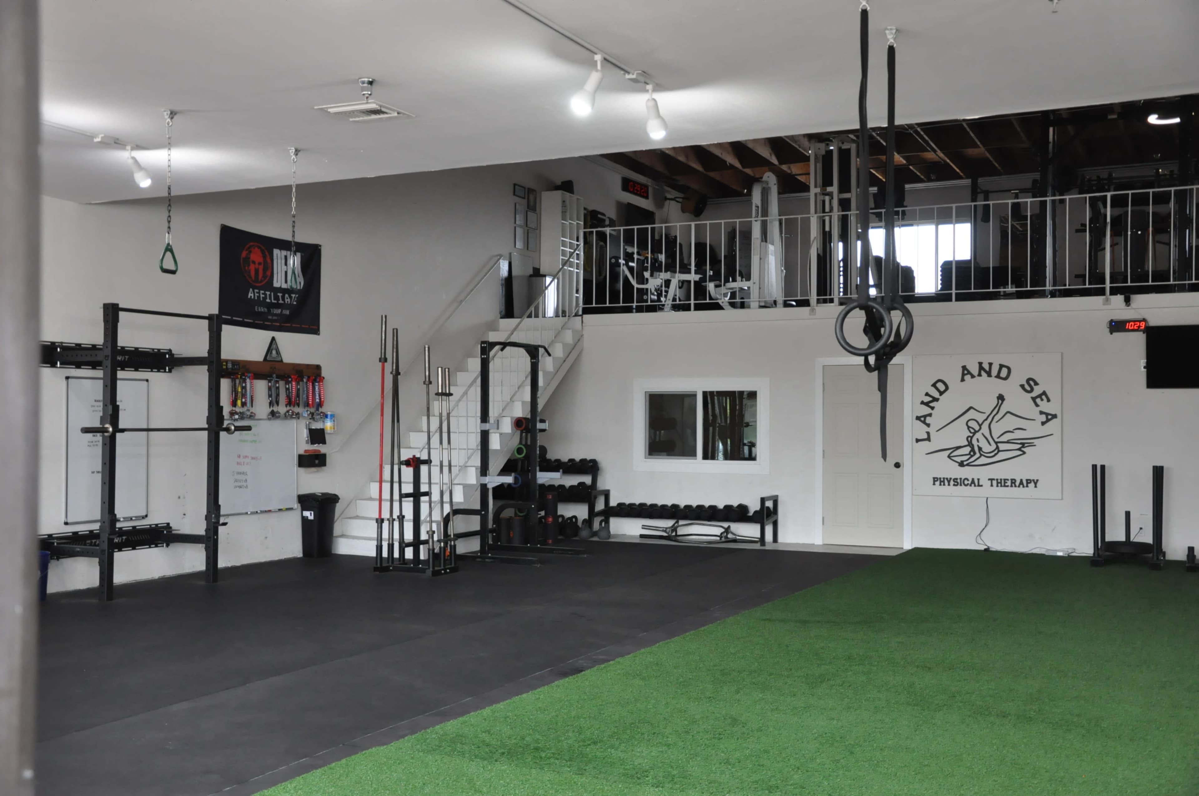 A spacious gym with a turf area, various exercise equipment, and a wall featuring the name "Land and Sea Physical Therapy."