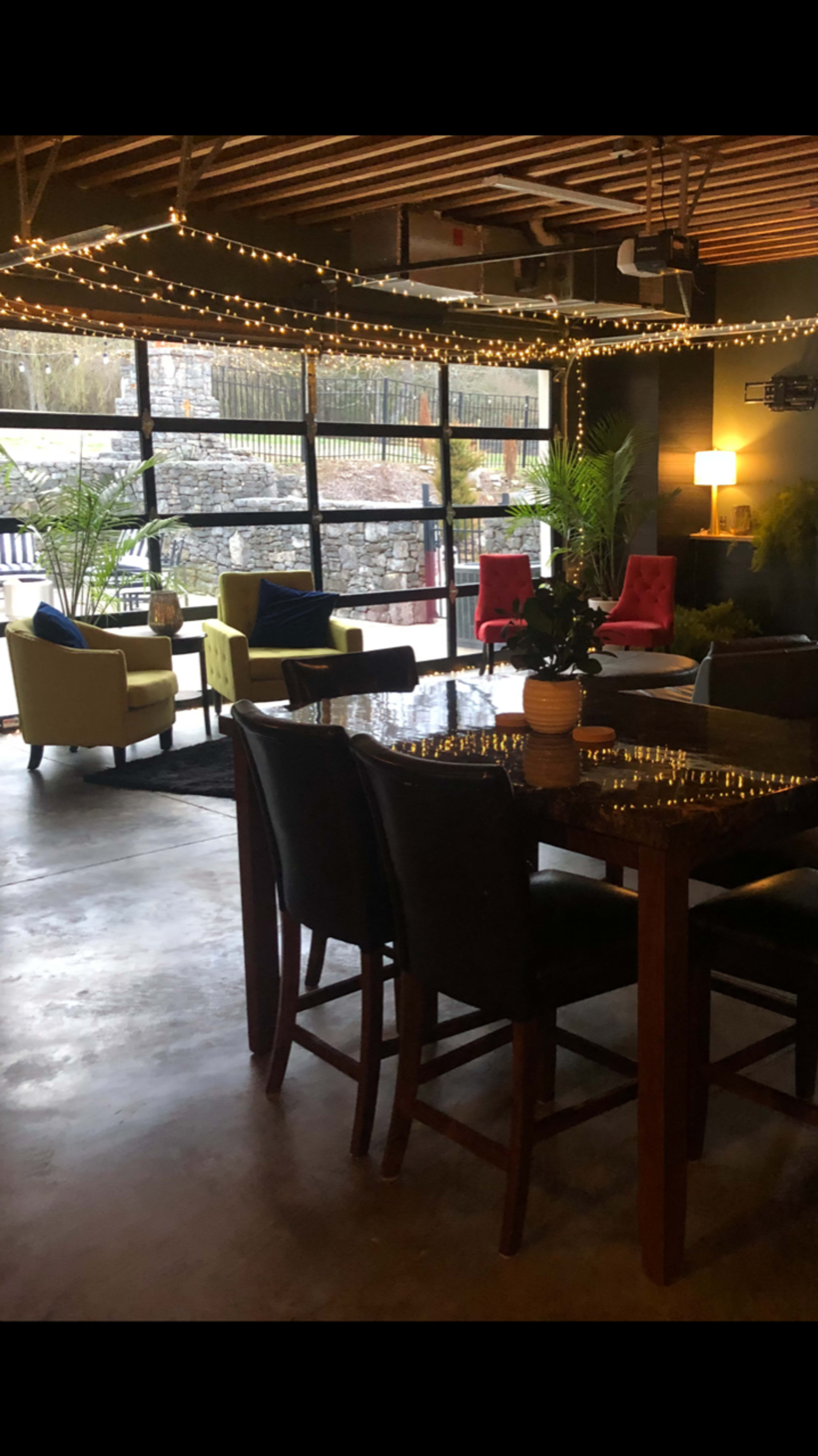 A cozy indoor seating area featuring a dining table with chairs, decorated with string lights and potted plants, alongside various colorful armchairs.