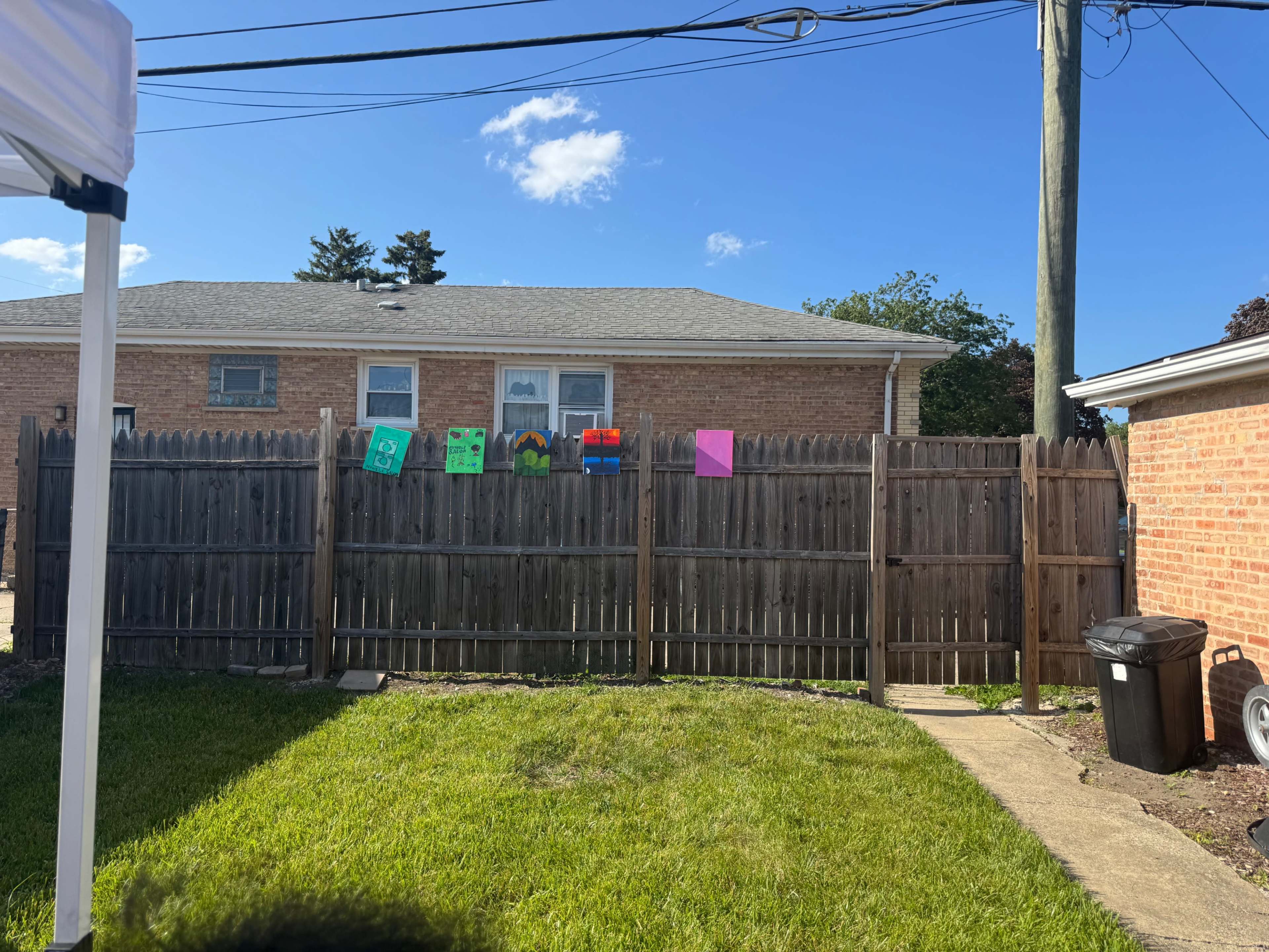 The image shows a wooden fence with a variety of colorful signs attached to it, set against a background of a house and a clear blue sky.