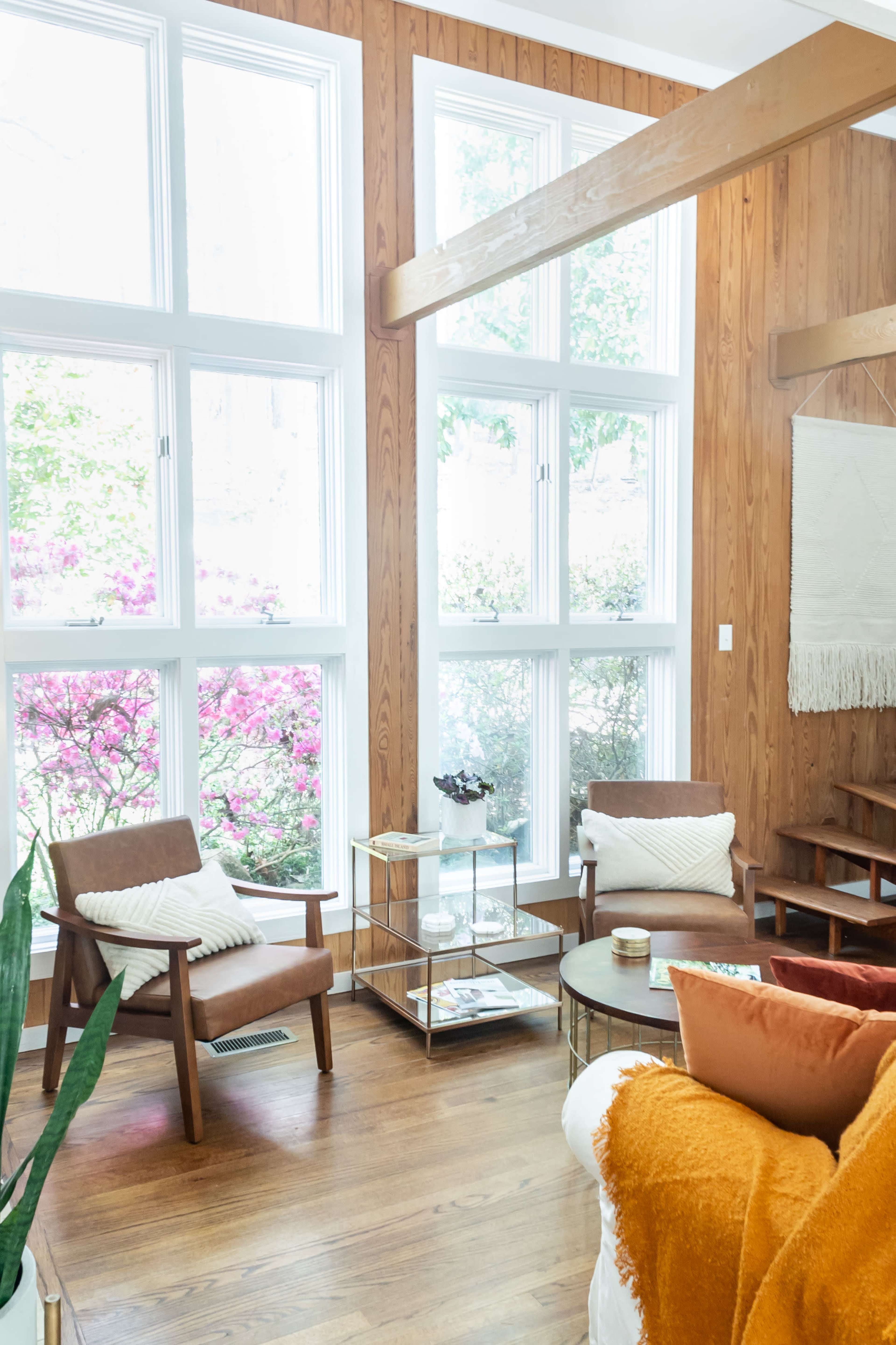 A cozy living room with large windows overlooking blooming pink flowers, featuring two wooden armchairs and a coffee table on a hardwood floor.