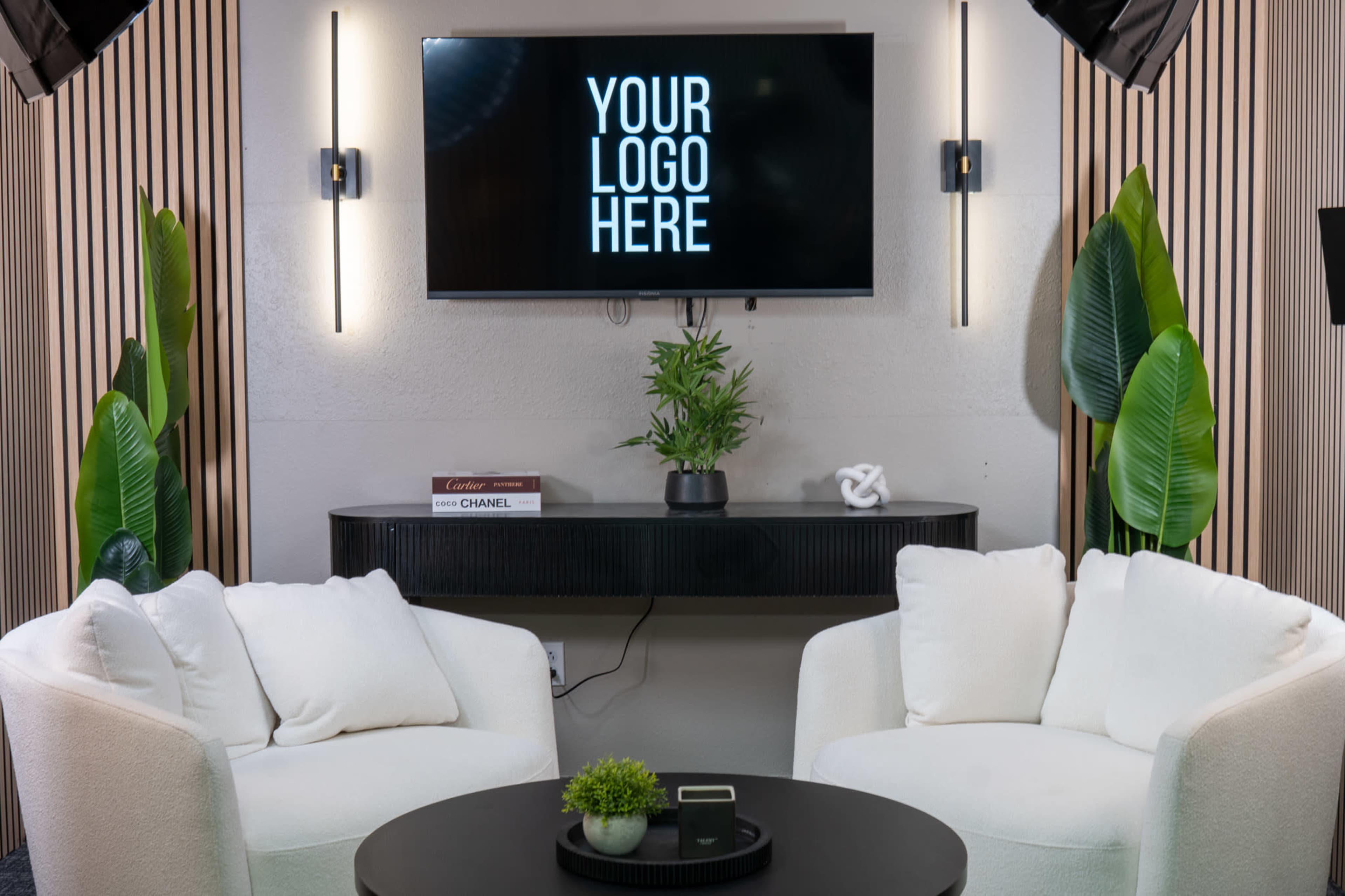 A modern studio setup features two white armchairs facing a circular black table, with a television displaying "YOUR LOGO HERE" and green plants in the background.