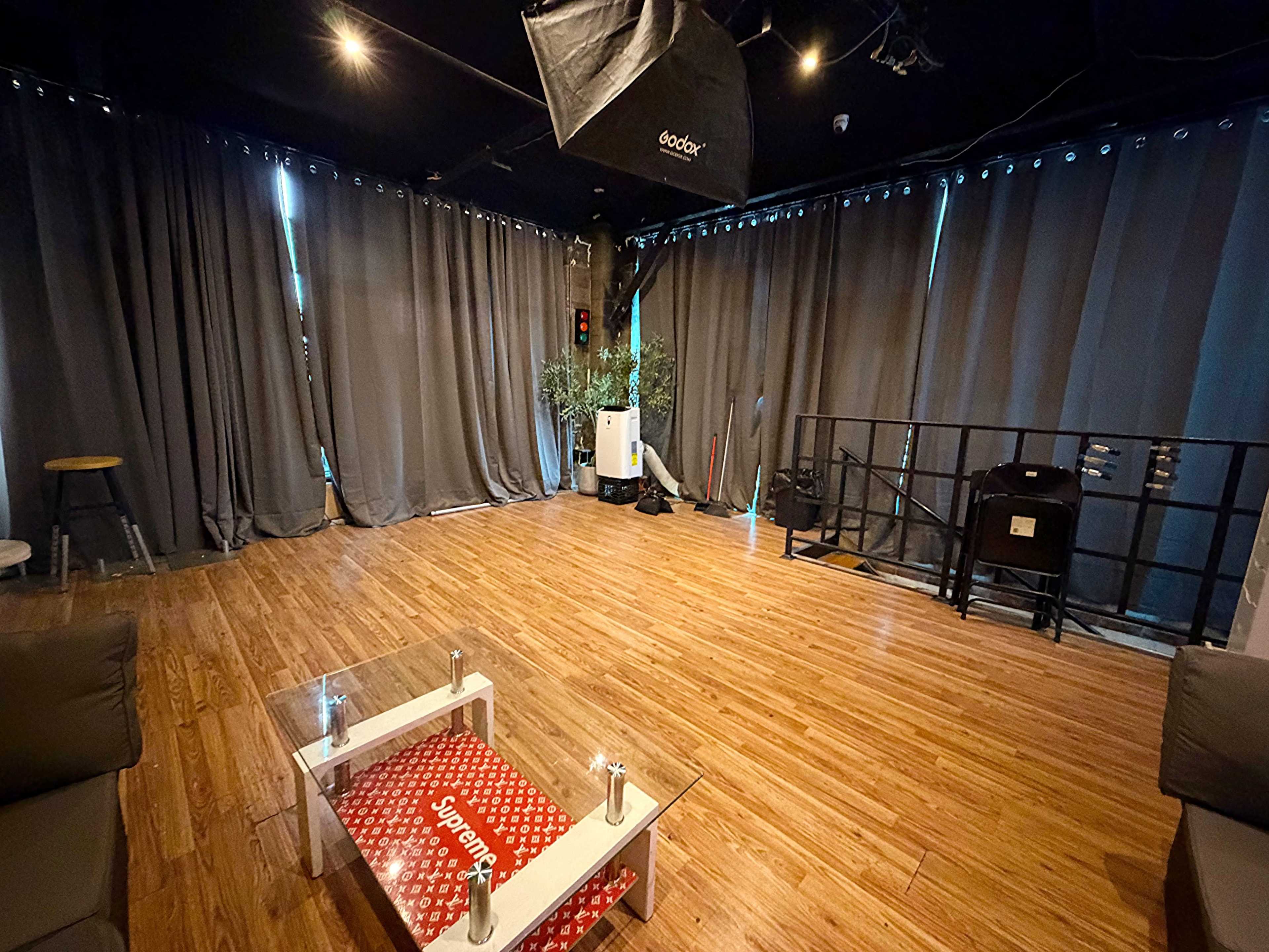 The image shows a spacious room with wooden flooring, surrounded by dark curtains, featuring a low glass table and a few furniture pieces.
