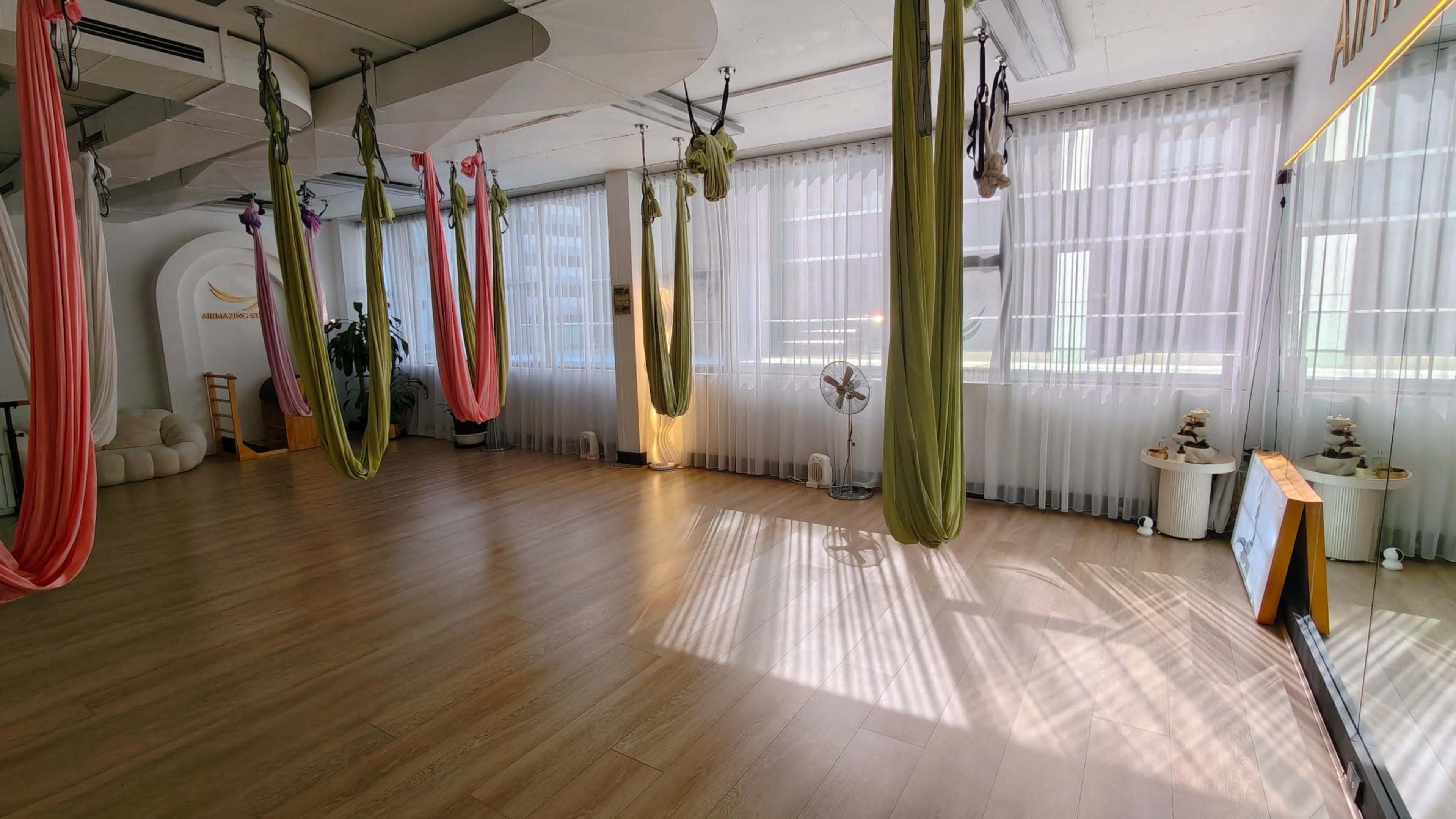 The image shows a spacious yoga studio with suspended hammocks in various colors, large windows with sheer curtains, and wooden flooring.