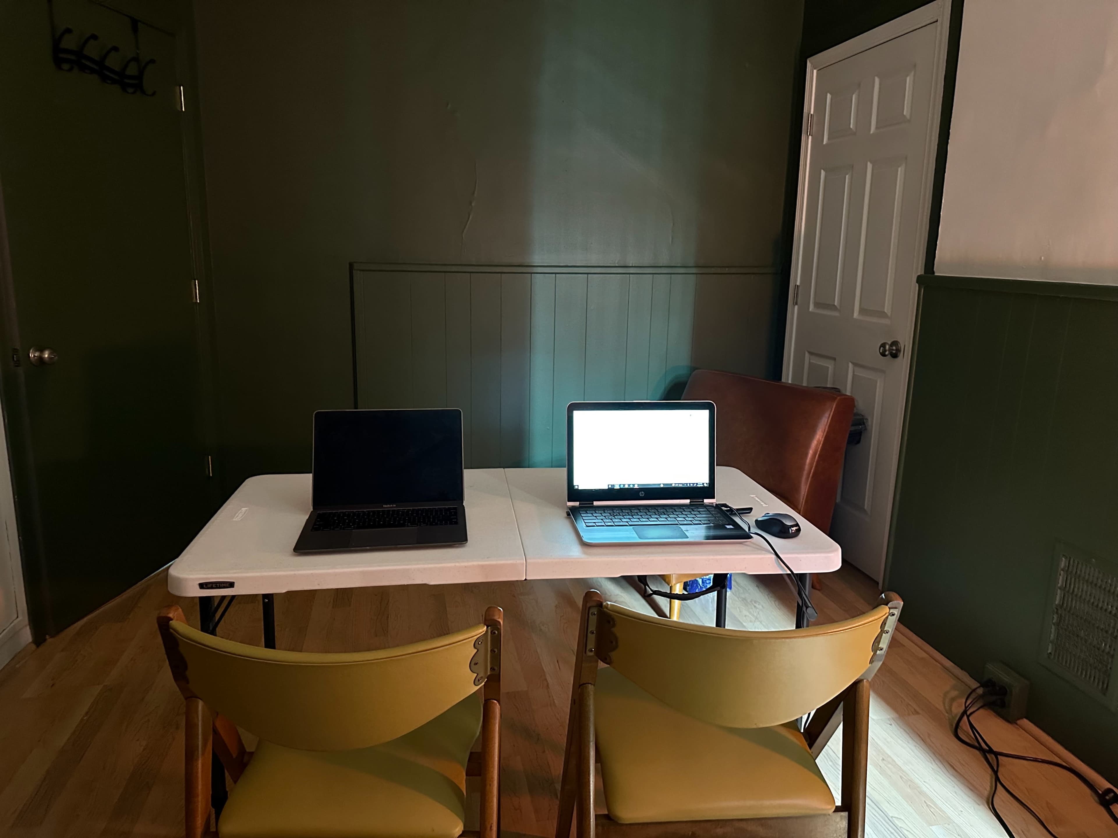 A small room features a table with two laptops and two chairs, against a green wall with a door in the background.