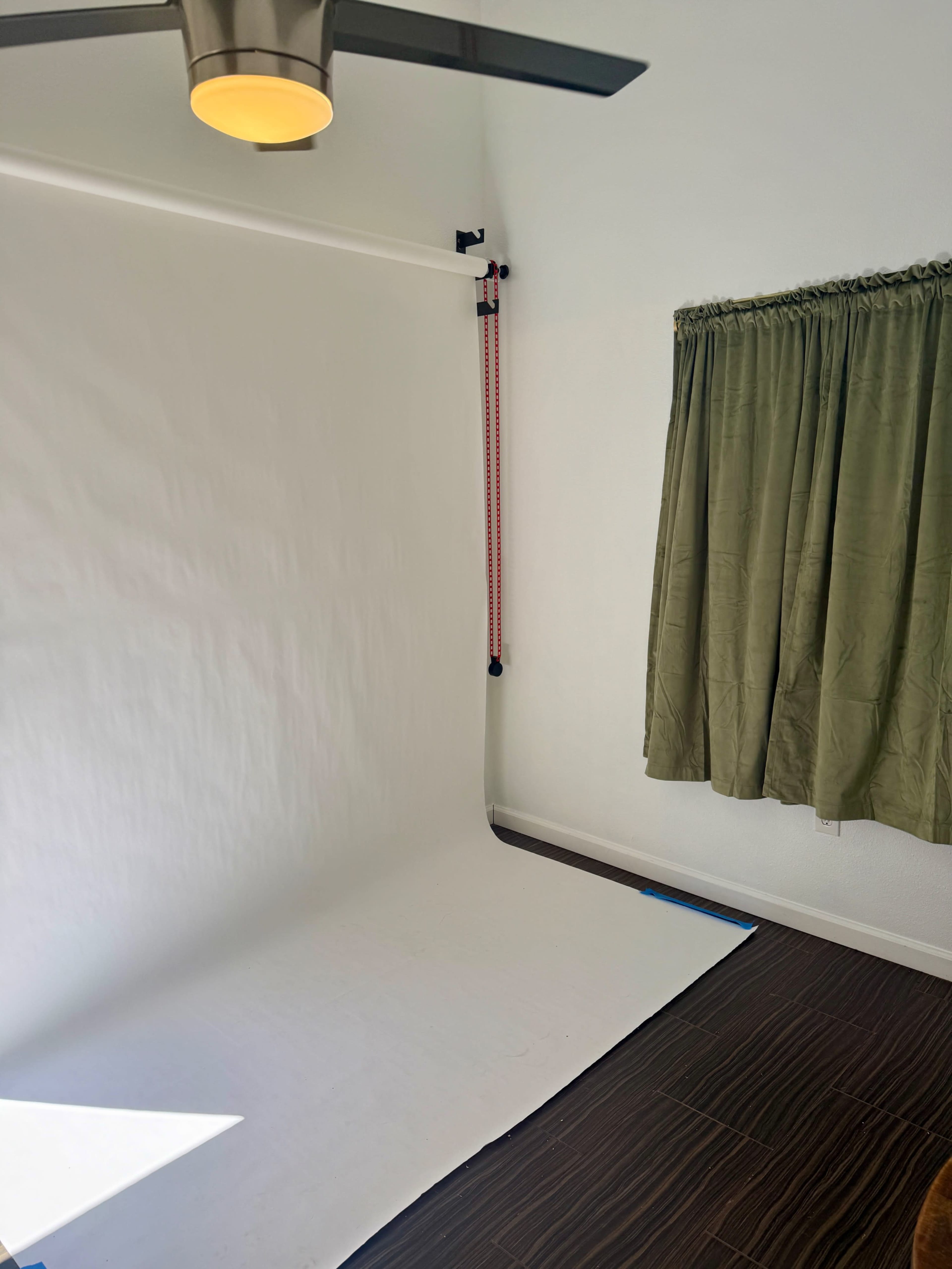 The image shows a empty photography studio with a white backdrop, a ceiling fan, and green curtains on the window.