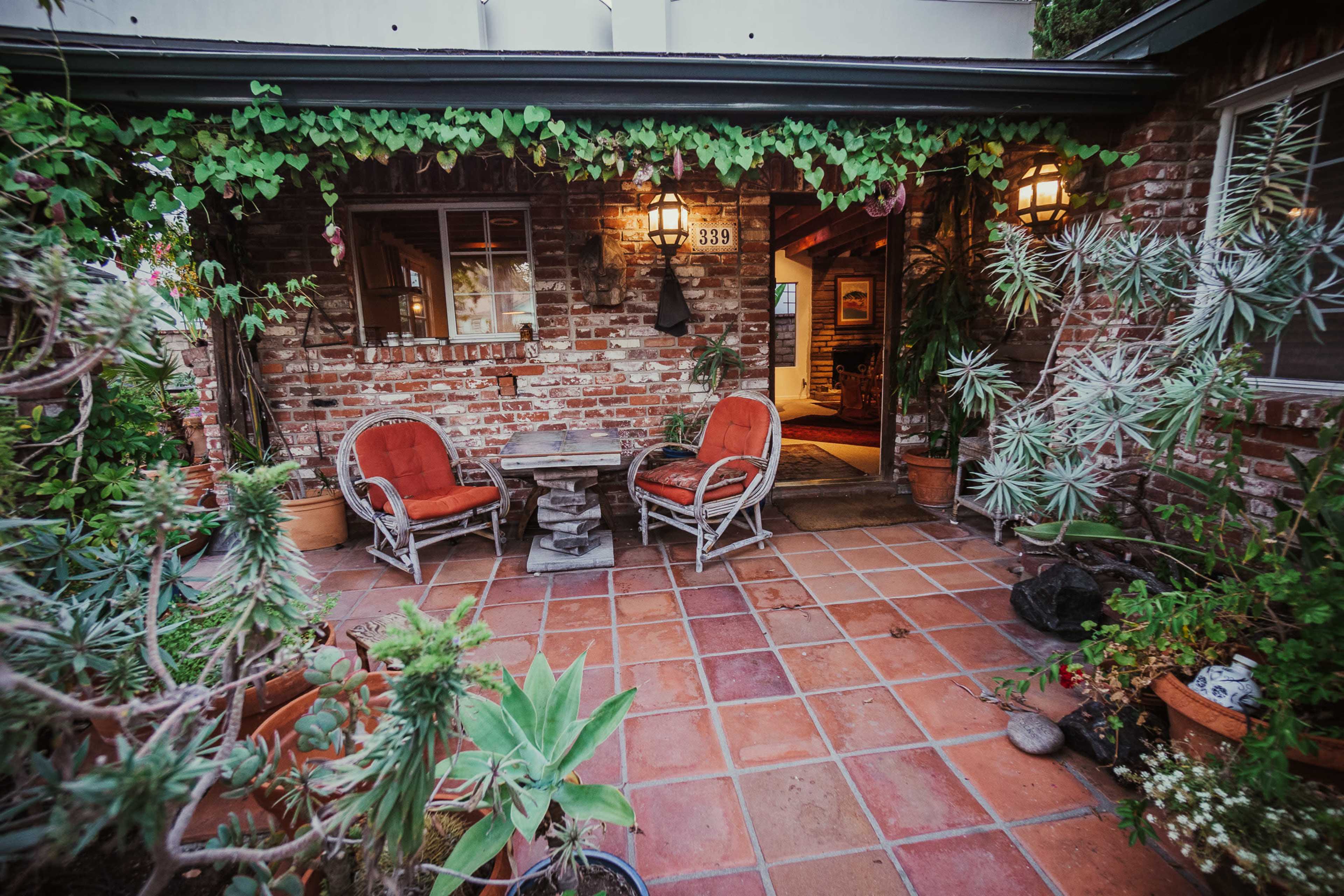 A brick patio area features a small table with two red chairs surrounded by potted plants and vines.