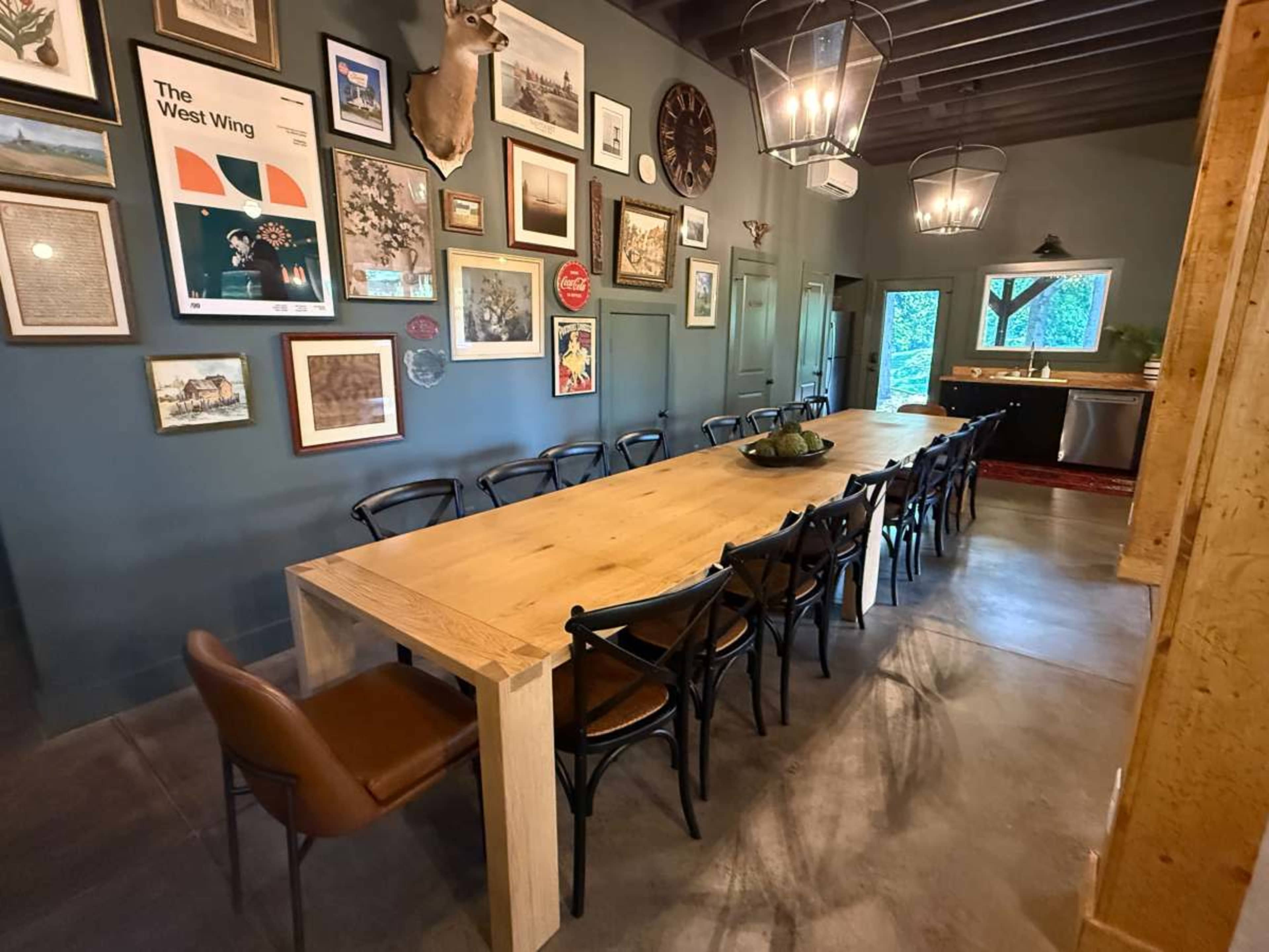 A long wooden dining table with black chairs is set against a dark wall adorned with various framed artworks and photographs.