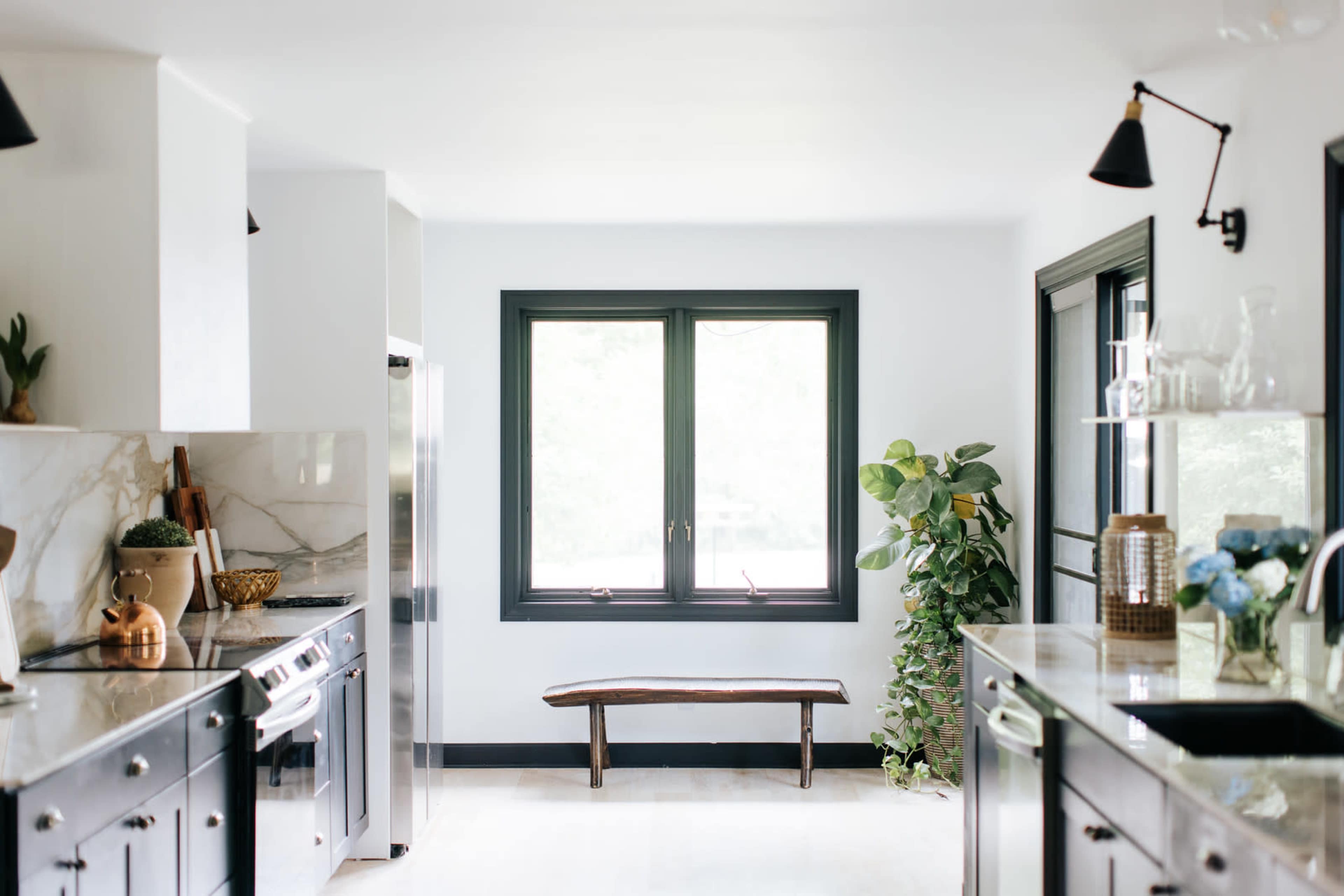 A modern kitchen features black cabinetry, marble countertops, and a wooden bench placed in front of a large window with greenery outside.
