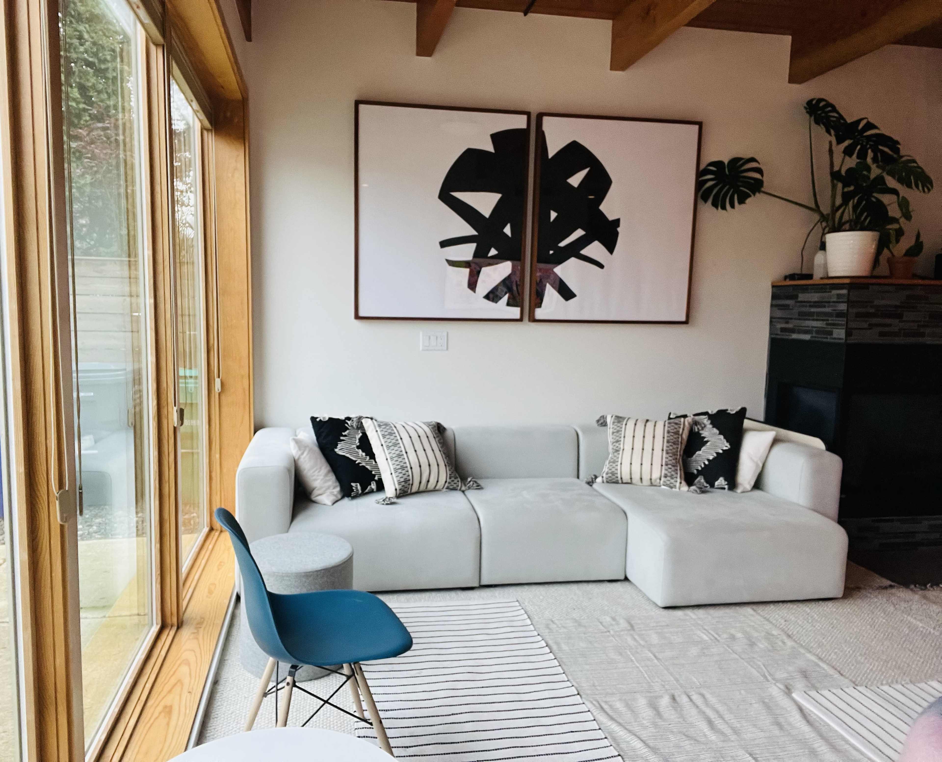 A modern living room features a light-colored sectional sofa, two abstract framed artworks on the wall, a small round side table, and a blue chair beside large windows that provide natural light.