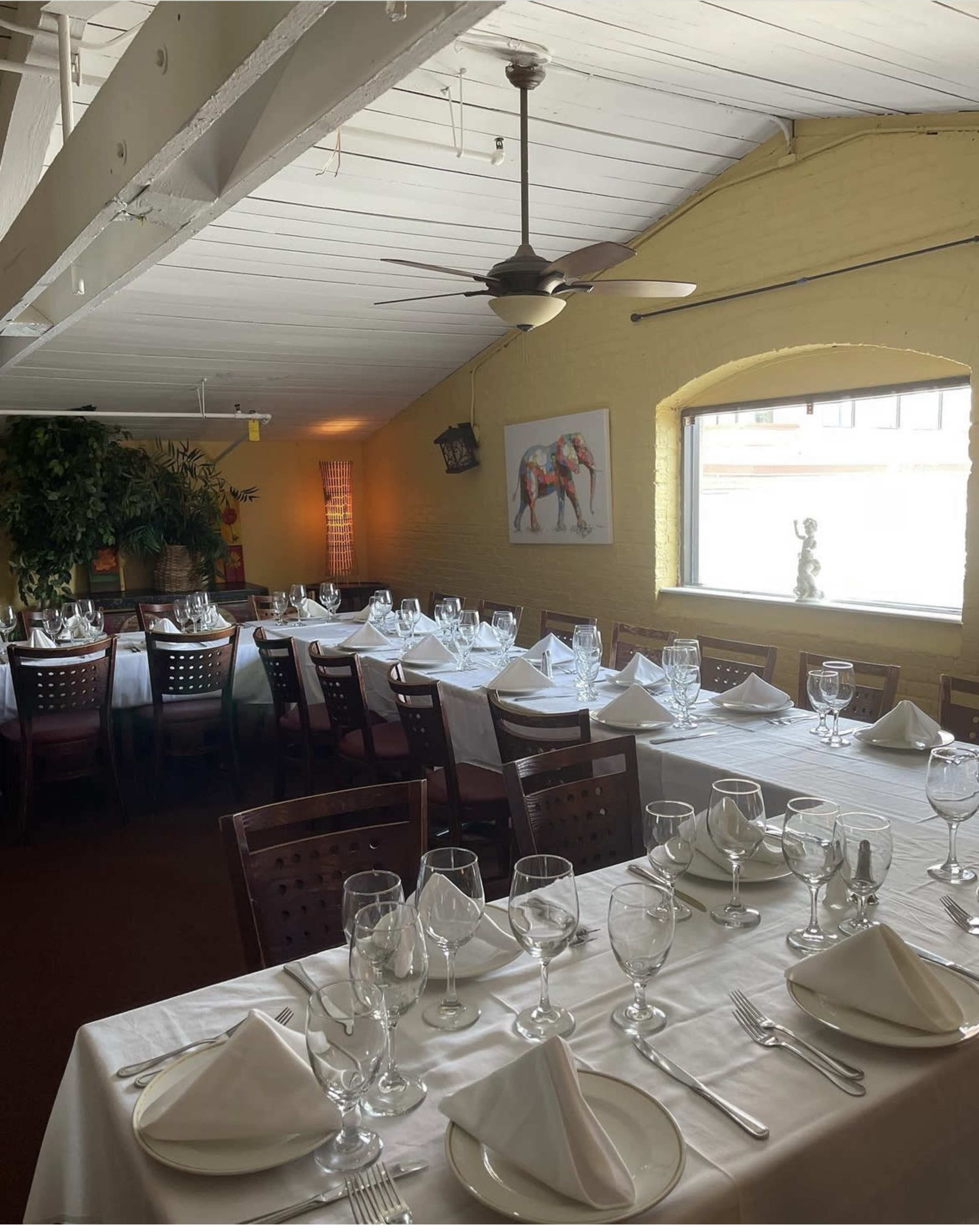 A well-prepared dining area with neatly arranged tables, each set with white tablecloths, glassware, and silverware, under a ceiling fan.