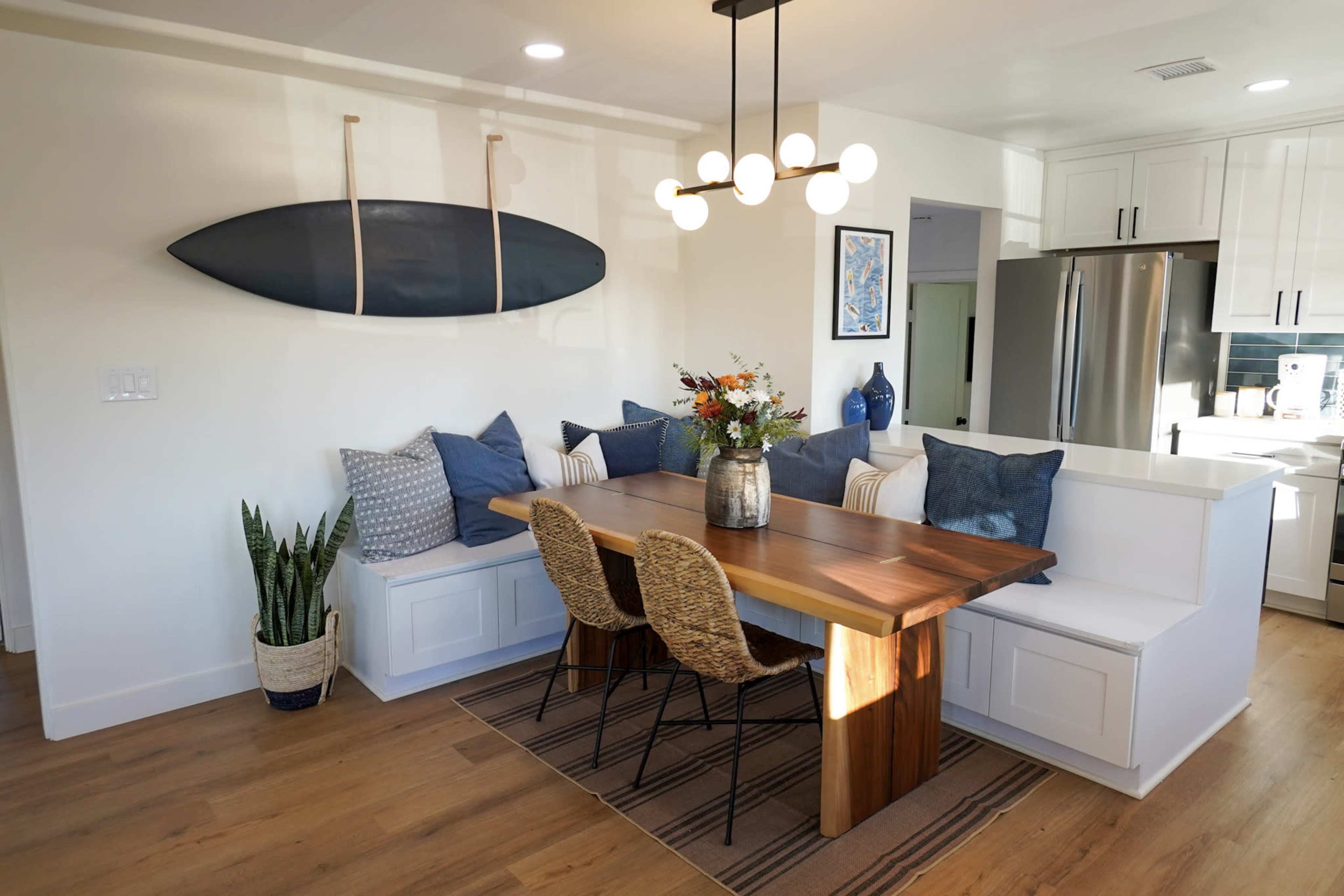 A dining area features a wooden table surrounded by woven chairs, with a built-in bench adorned with blue pillows and a surfboard mounted above.