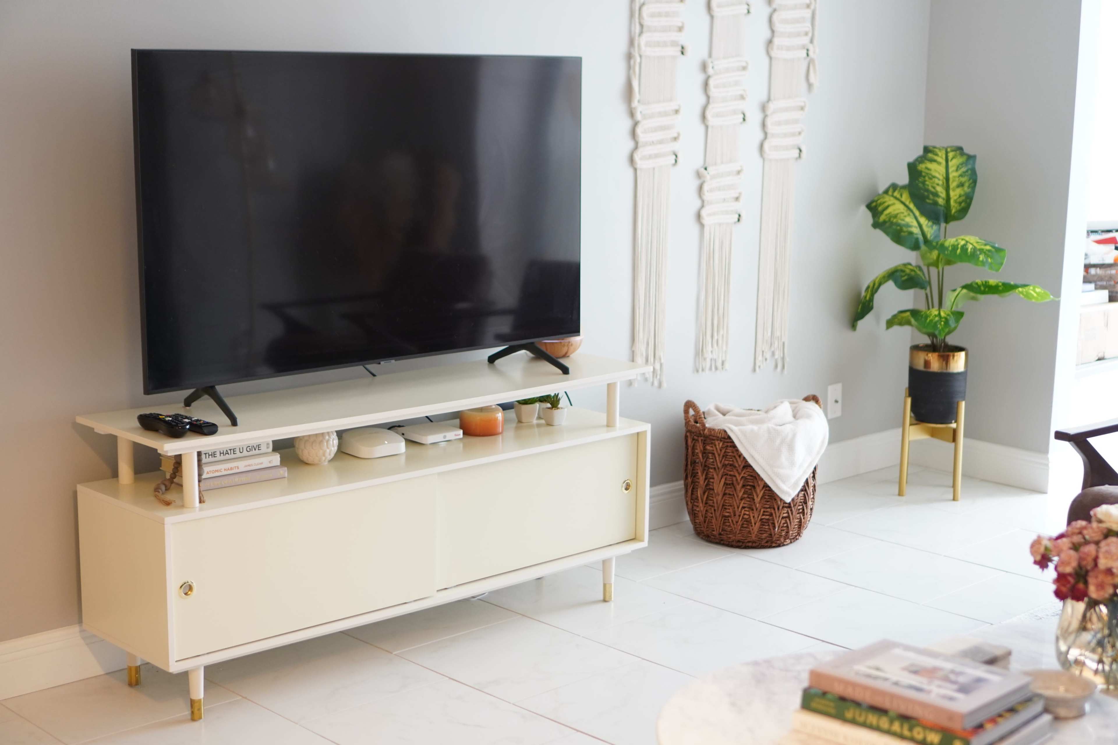 The scene features a modern living room with a large flat-screen TV mounted above a light-colored entertainment unit, accompanied by a decorative basket and a potted plant.
