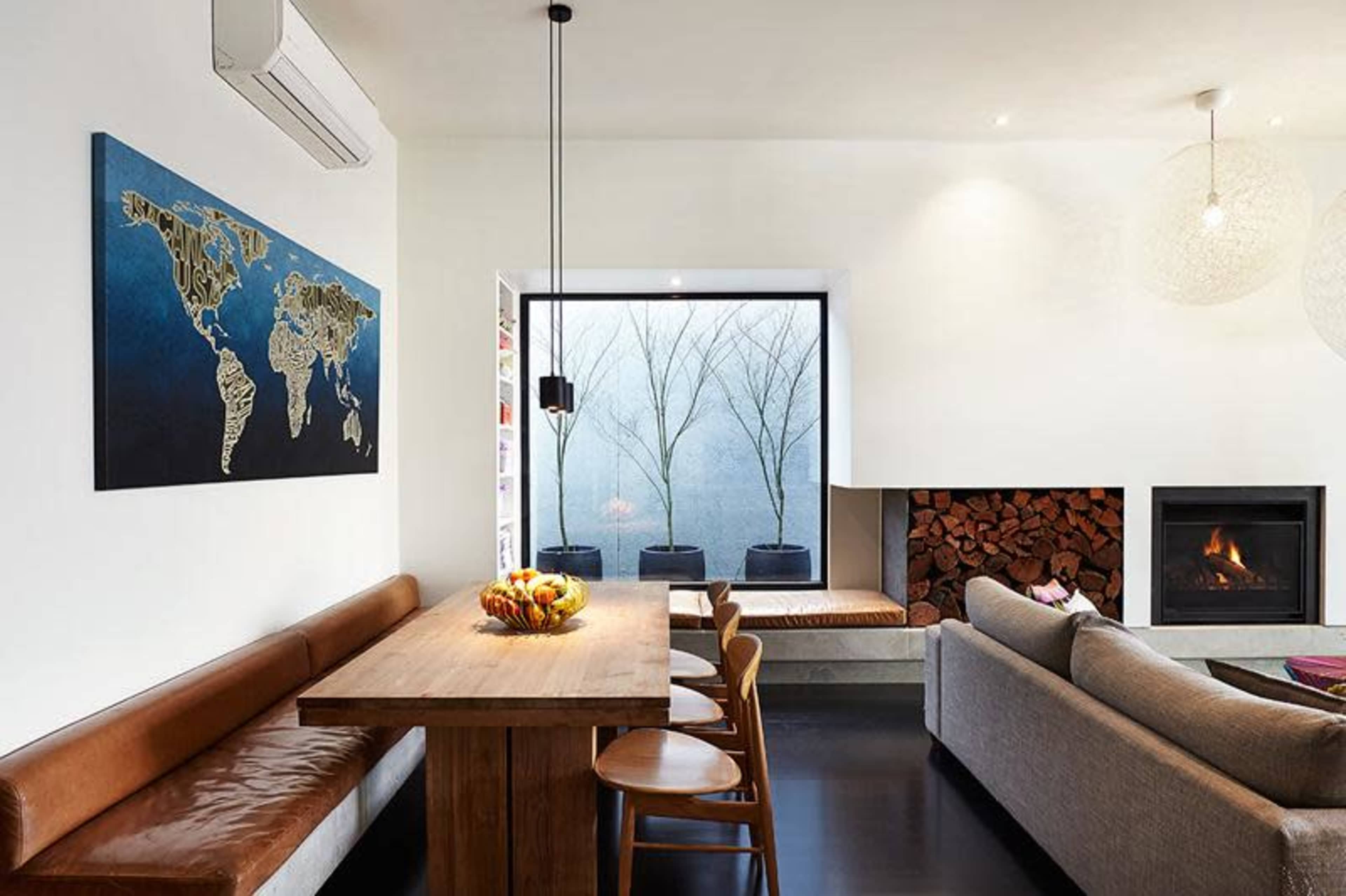 A spacious living room features a wooden dining table with seating, a large window view of bare trees, and a wall-mounted fireplace.