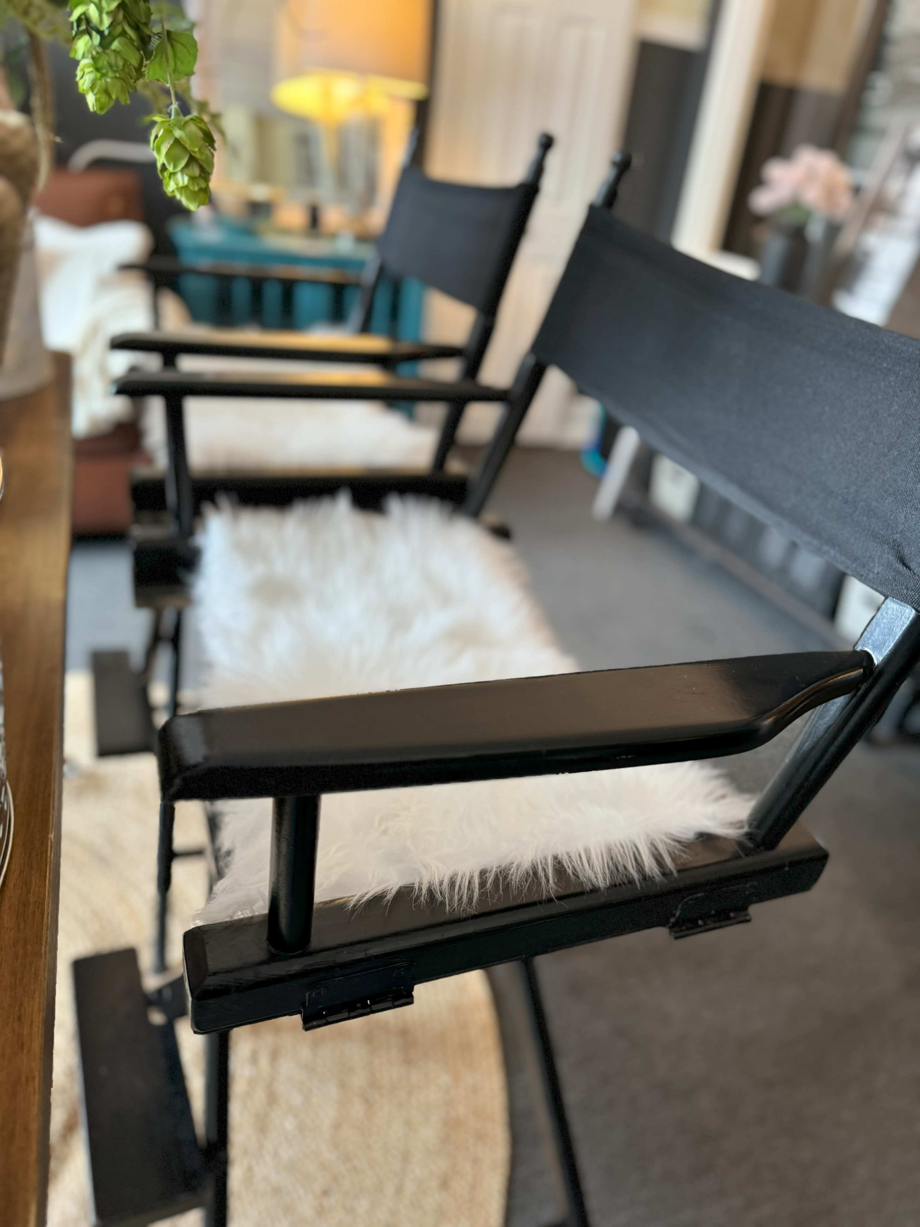 Two black chairs with armrests, each featuring a white fur cushion, are positioned beside a wooden table in a room with decorative elements.