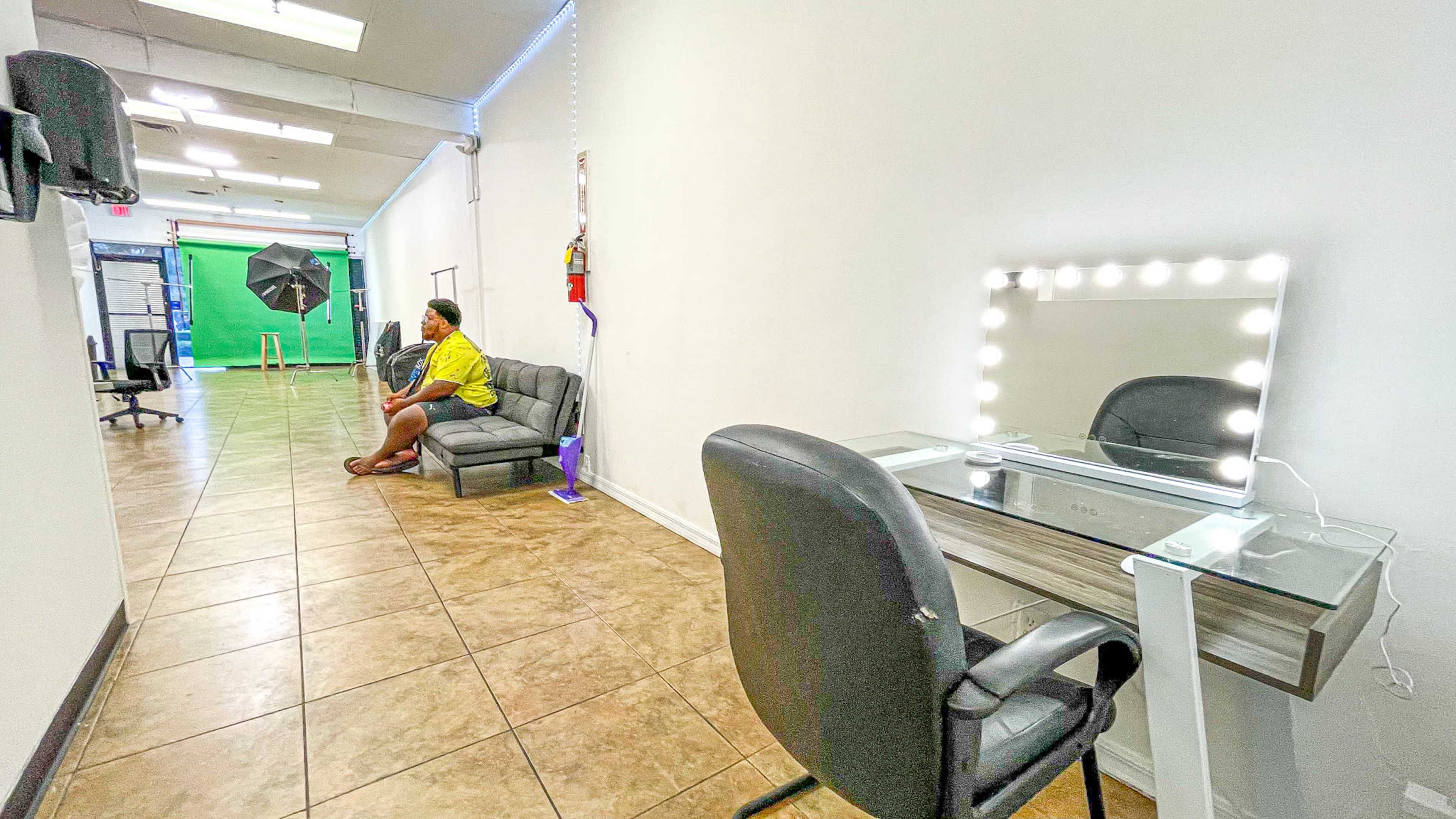 A woman in a yellow outfit sits on a couch in a bright, sparsely furnished studio hallway with a makeup vanity and backdrop setup.