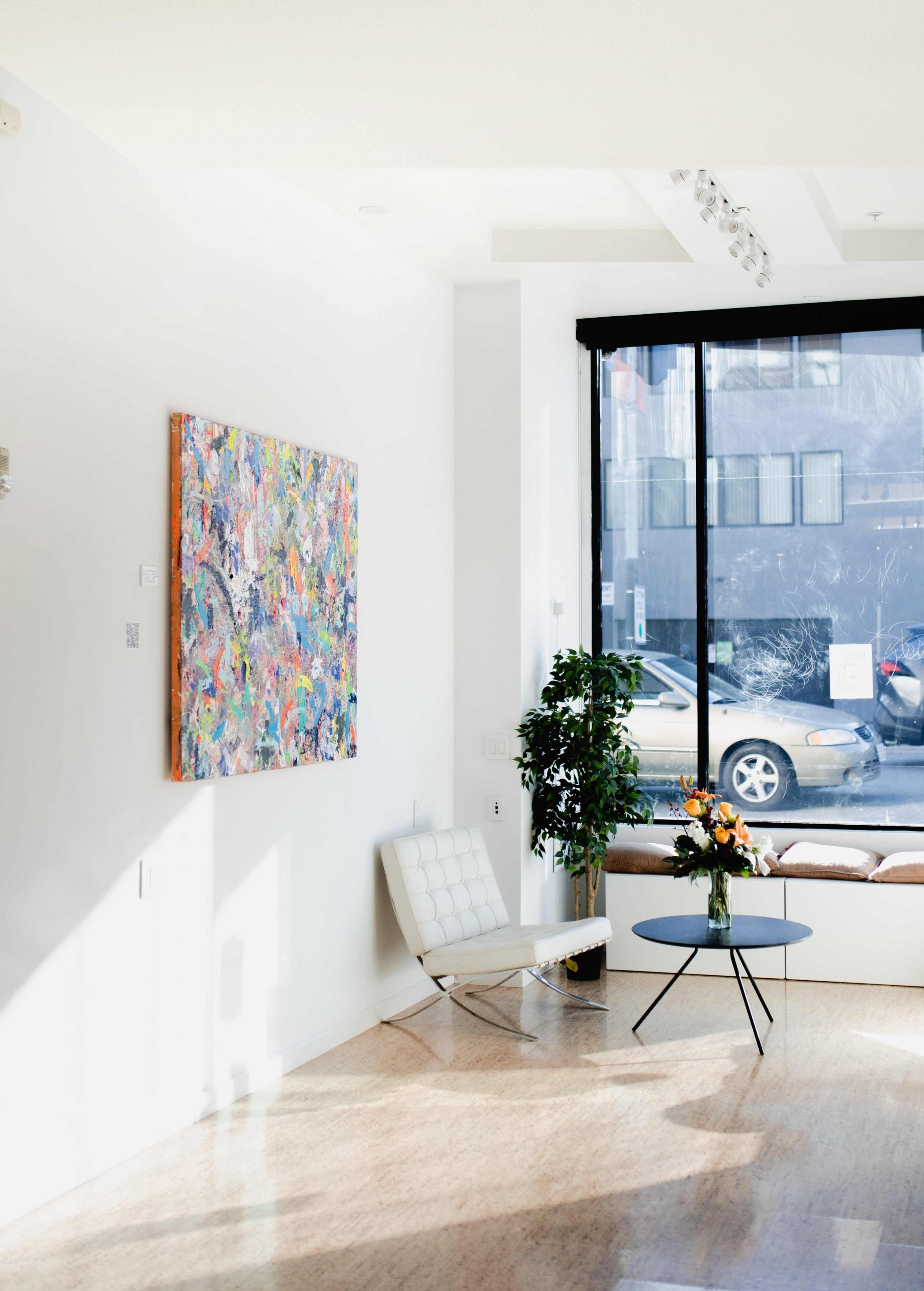 A bright, modern interior with a colorful painting on the wall, a sleek white chair, a small round table with flowers, and a large window overlooking a street.