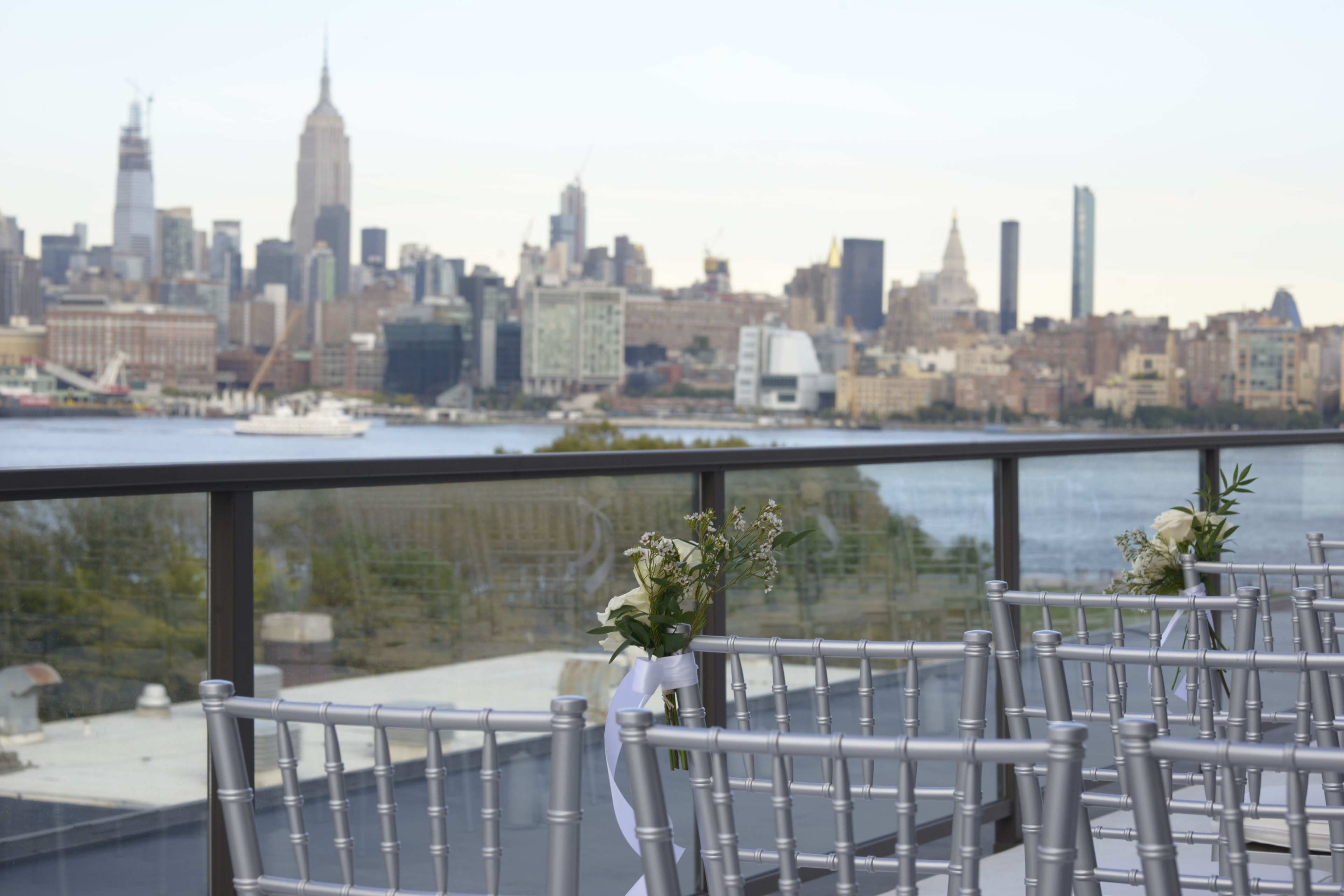 A silver chiavari chair line is set up on a balcony overlooking the New York City skyline, featuring the Empire State Building.