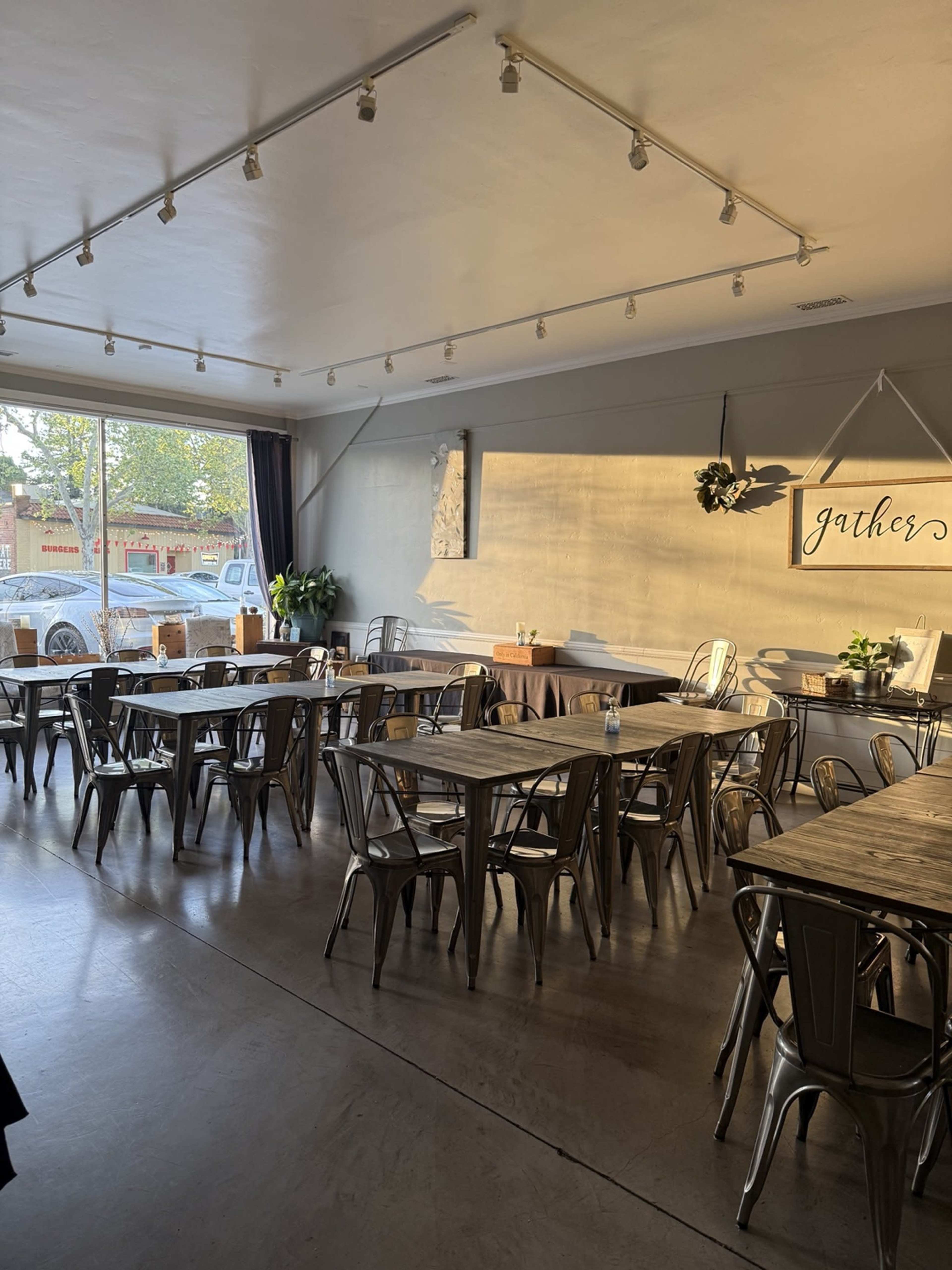 The image shows a modern, open restaurant interior with metal tables and chairs arranged in a spacious layout, featuring natural light streaming through large windows.