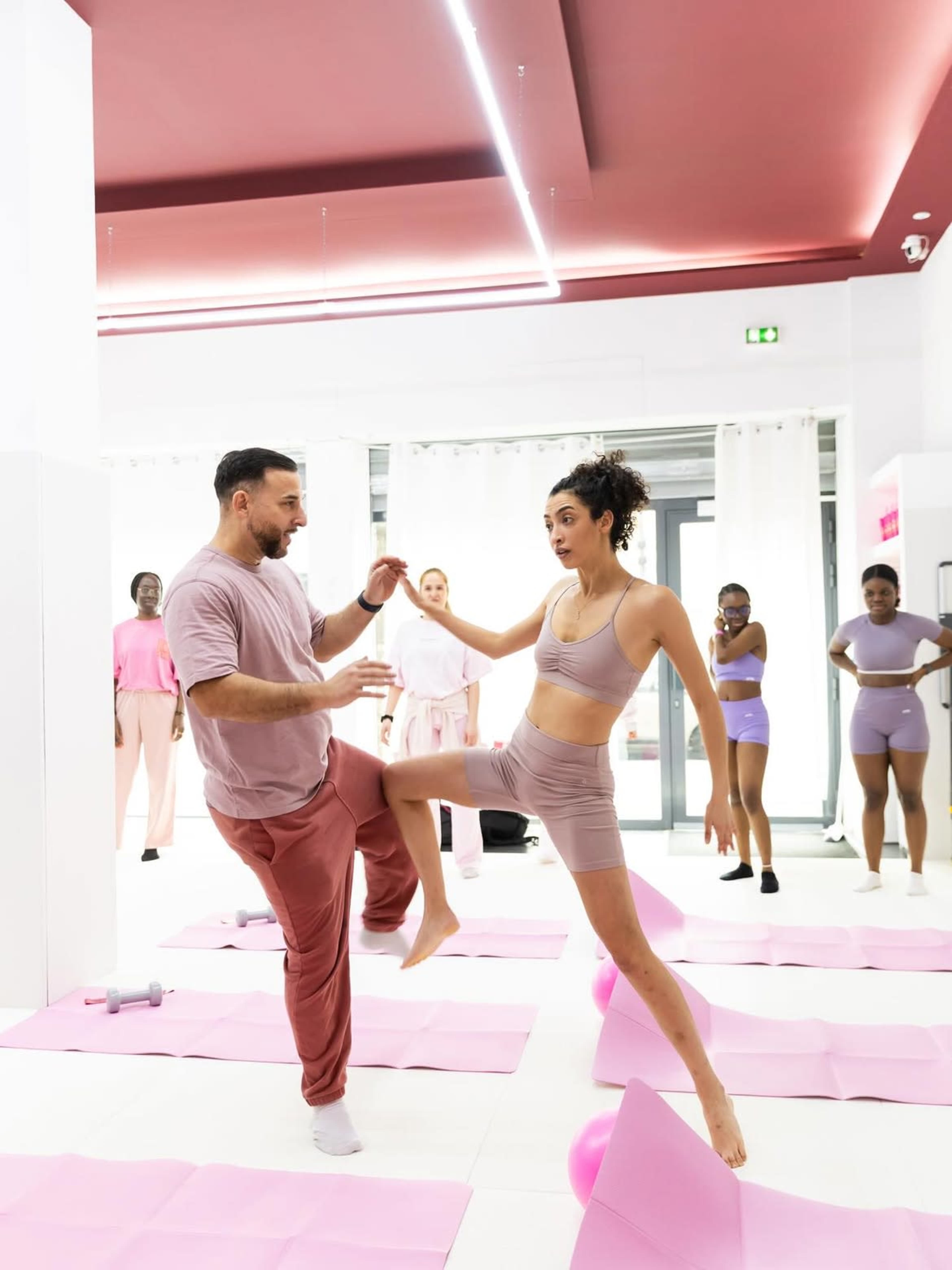 A group of people participates in a fitness class, with one instructor demonstrating a move while a participant mimics the action in a brightly lit studio.