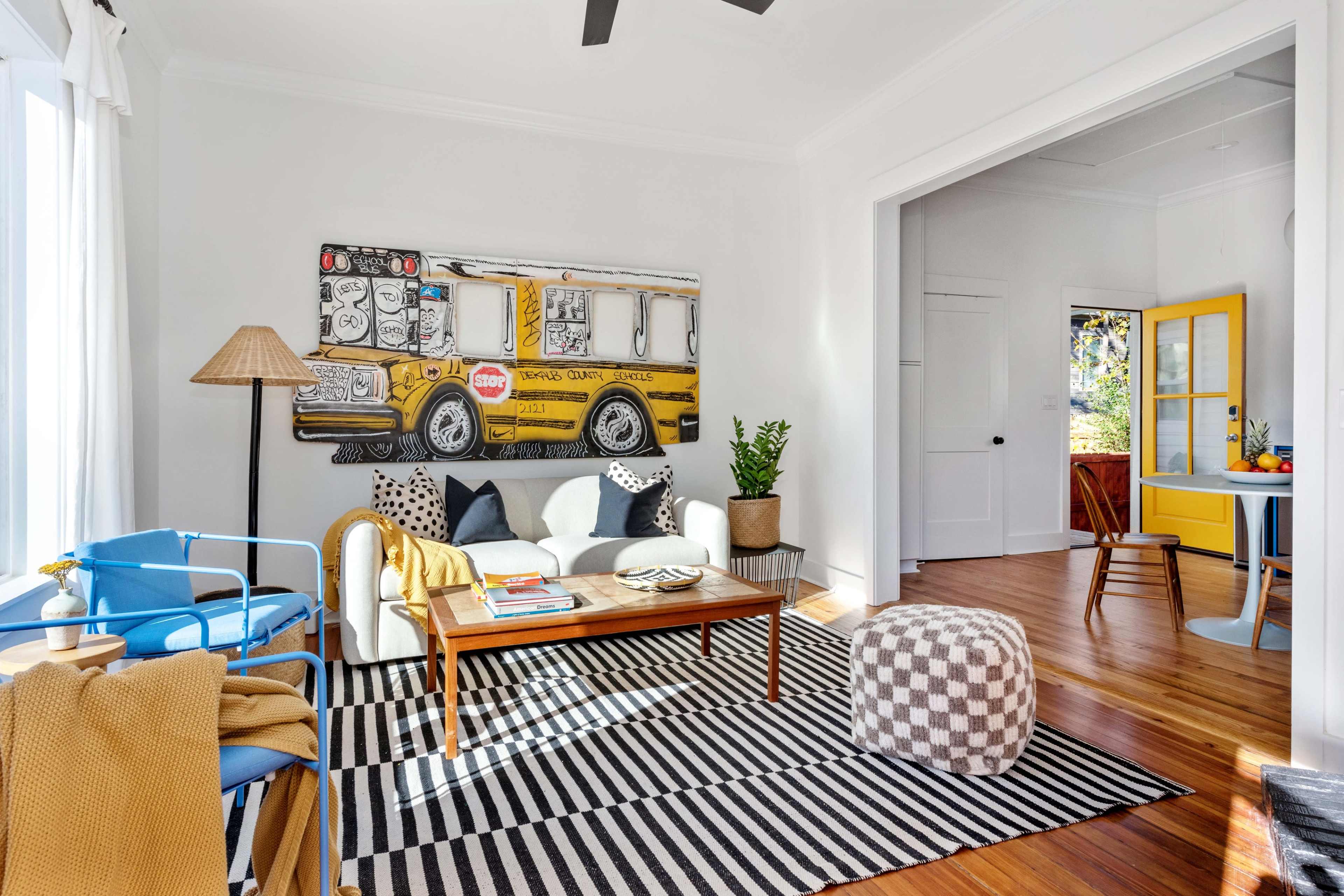 A bright living room features a large bus mural on the wall, a striped rug, a coffee table with books, and seating that includes a blue chair and a sofa adorned with decorative pillows.