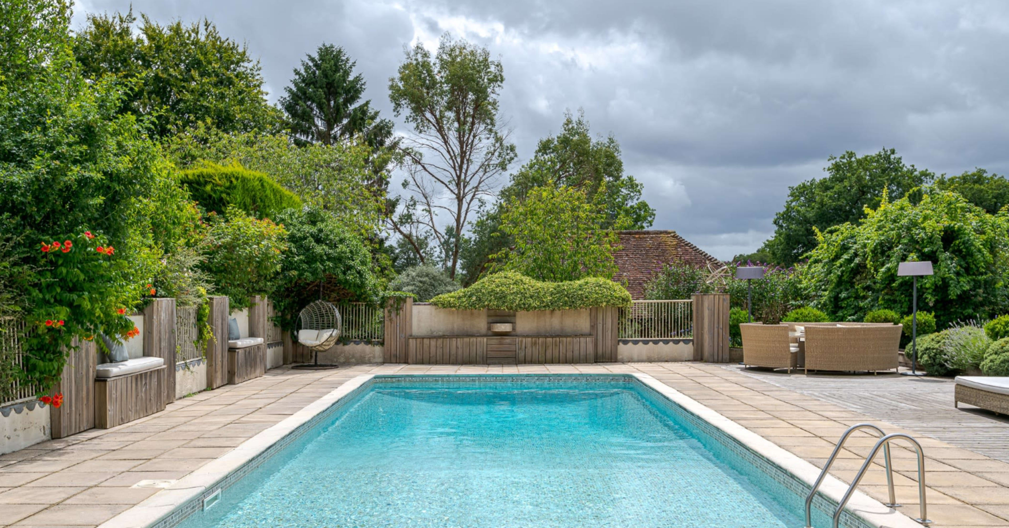 The image shows a rectangular swimming pool surrounded by greenery and patio seating under a cloudy sky.