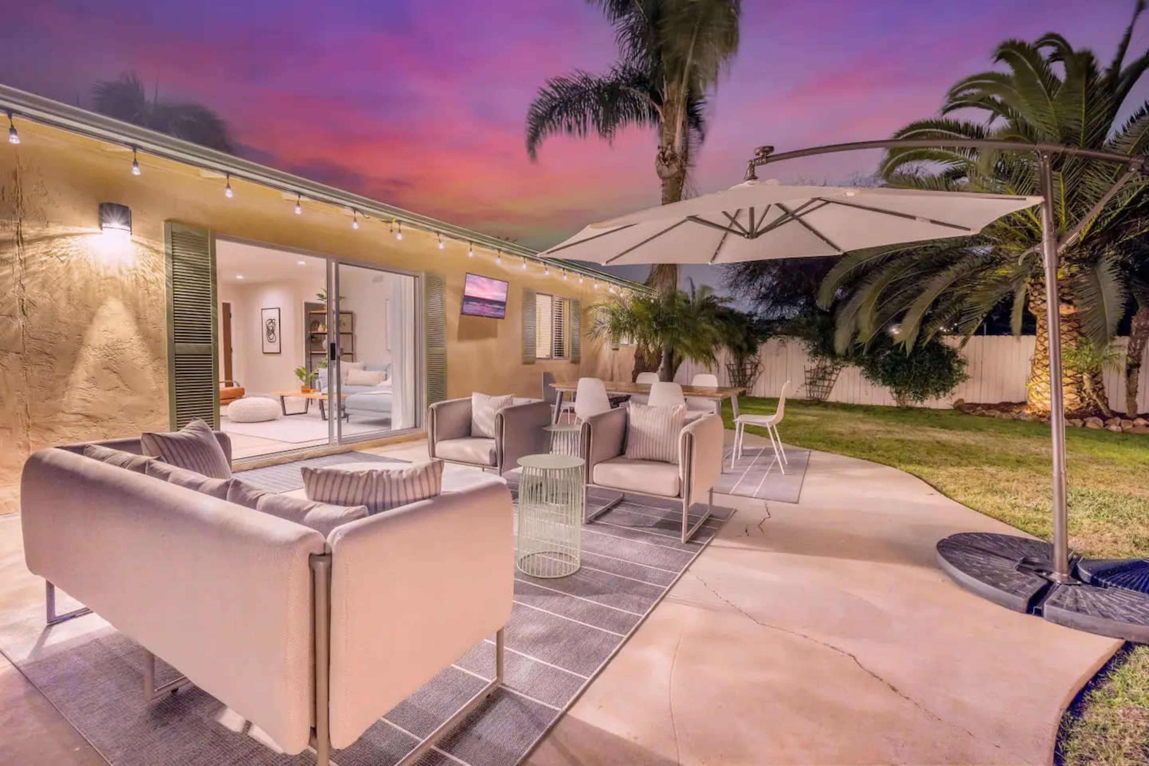 A cozy patio features modern seating, an umbrella, and vibrant palm trees under a colorful sunset sky.