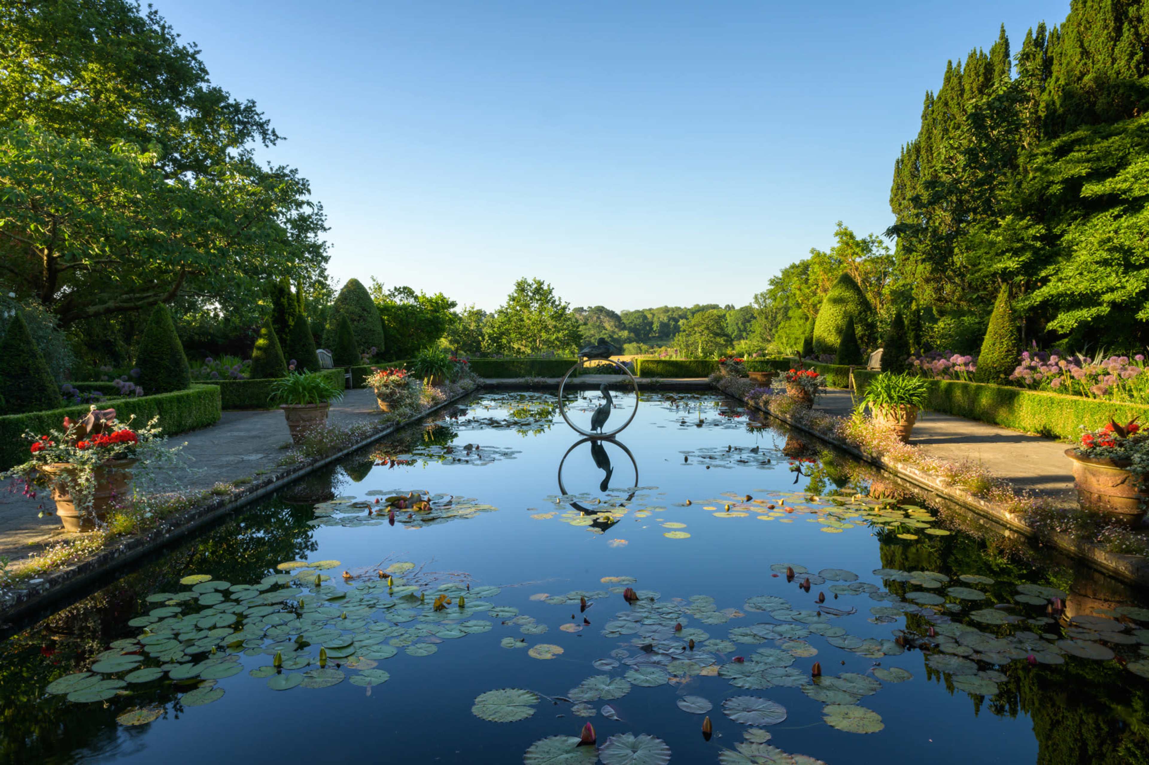 The scene features a tranquil garden with neatly arranged flower pots, a reflective pond adorned with lily pads, and a central sculpture framed by lush greenery.
