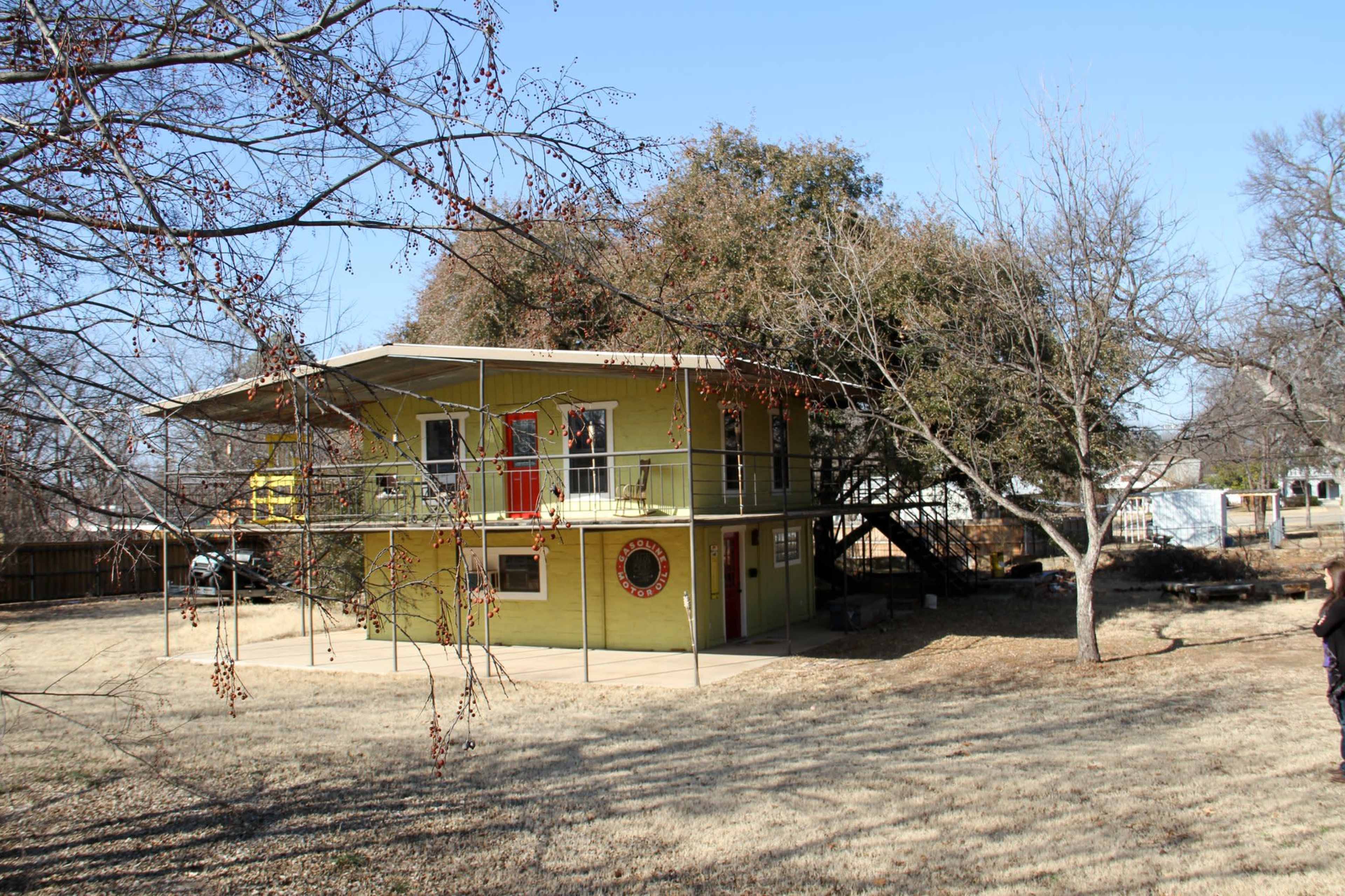 A two-story green house with a flat roof and a circular window is situated on a grassy lot surrounded by trees.