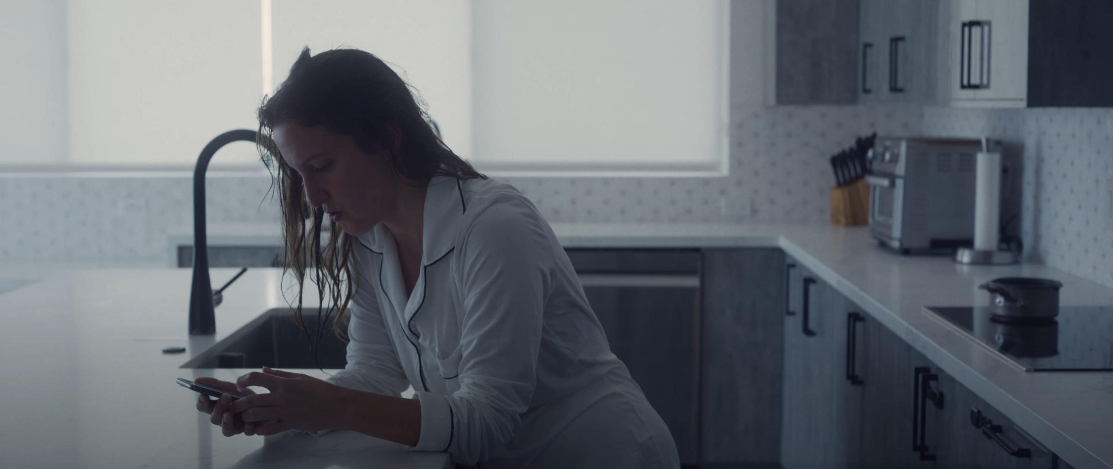 A woman with wet hair is leaning on a kitchen counter while looking at her phone.