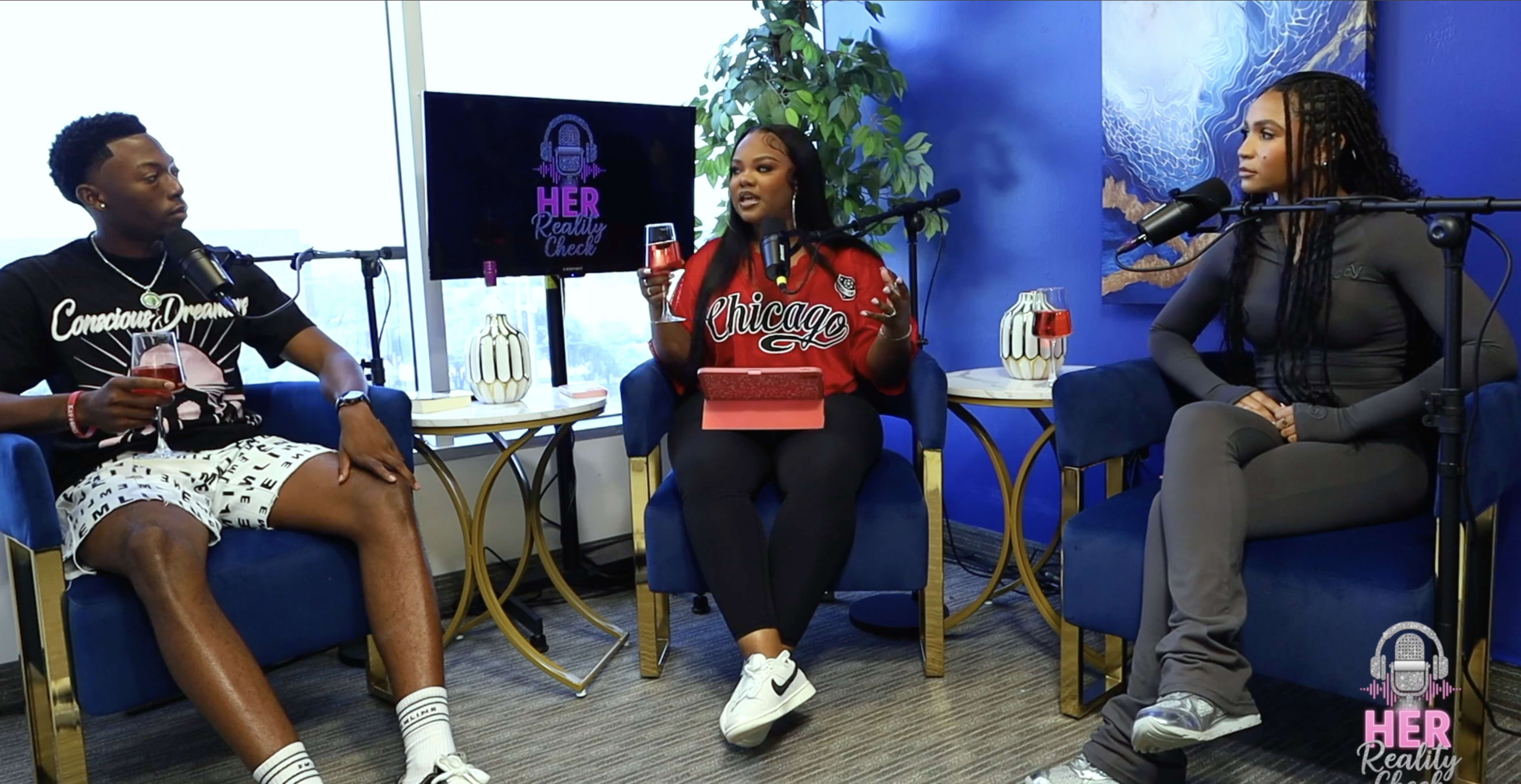 Three people are seated in modern blue chairs, engaging in conversation in a brightly lit room with windows, while the background features a blue wall and a decorative element.
