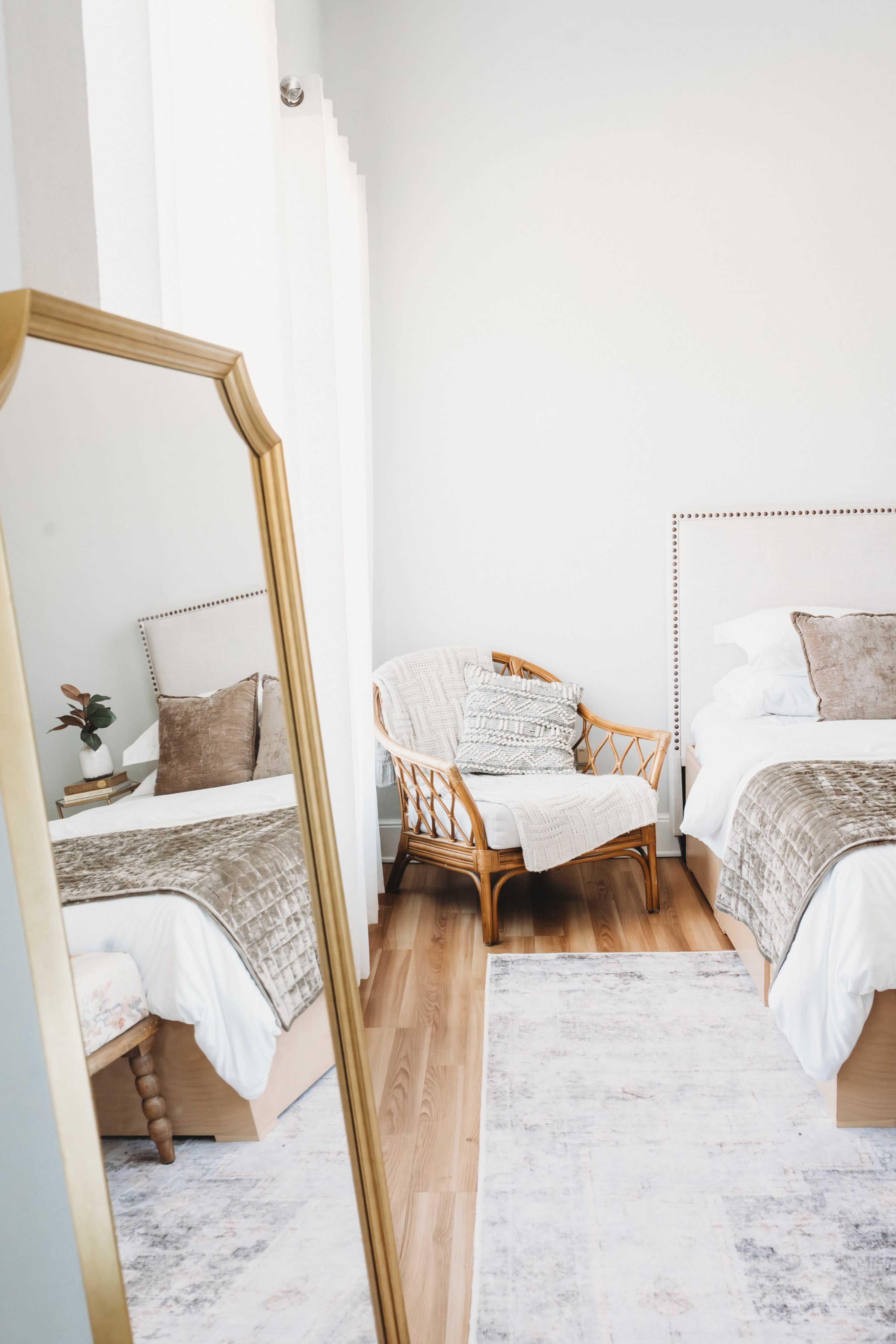 The image shows a bright bedroom featuring two beds, a decorative mirror, a rattan chair, and soft furnishings.
