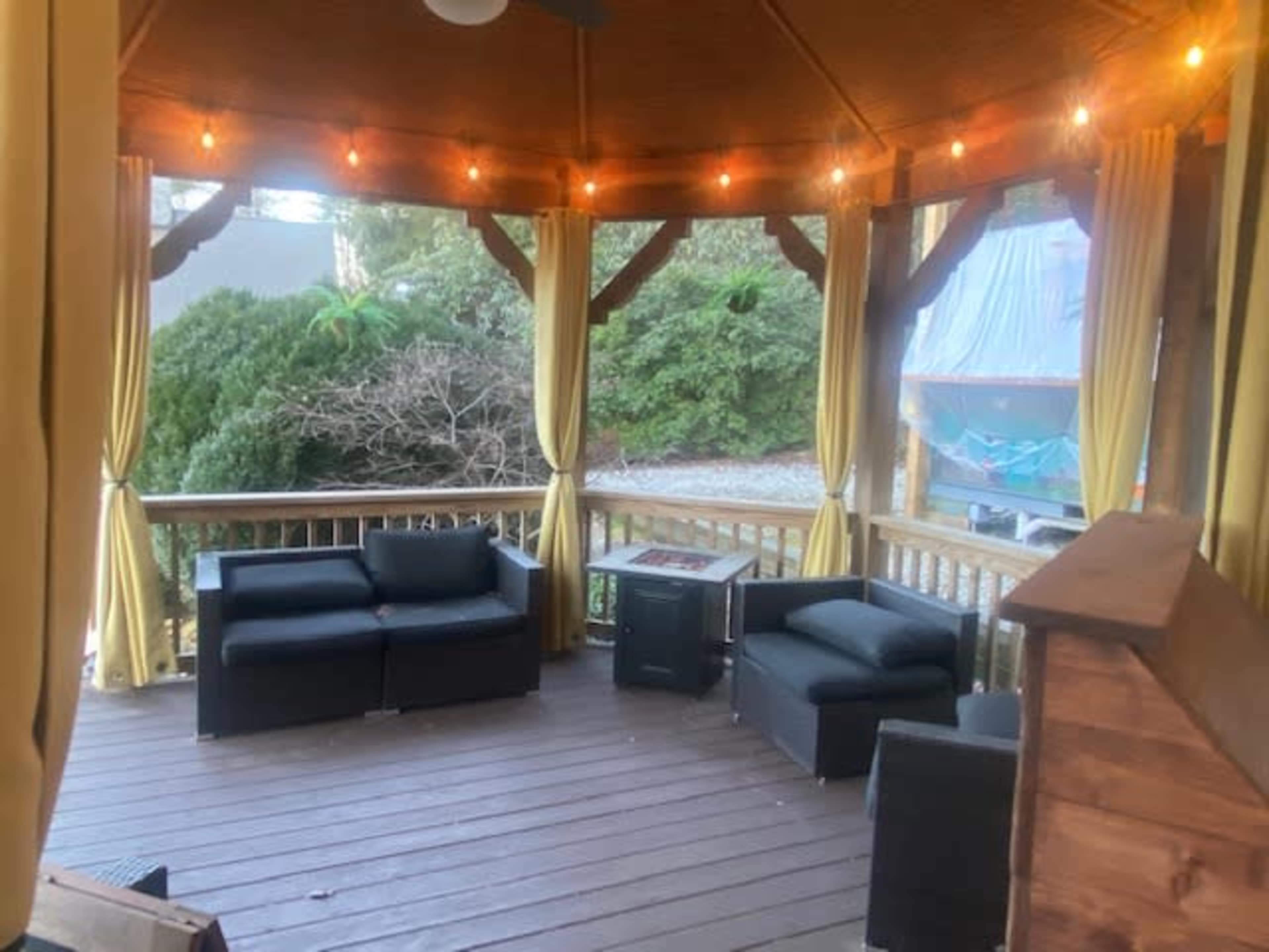 A gazebo with a wooden floor, featuring a seating arrangement of black couches and a fire pit, surrounded by yellow curtains and outdoor lighting.