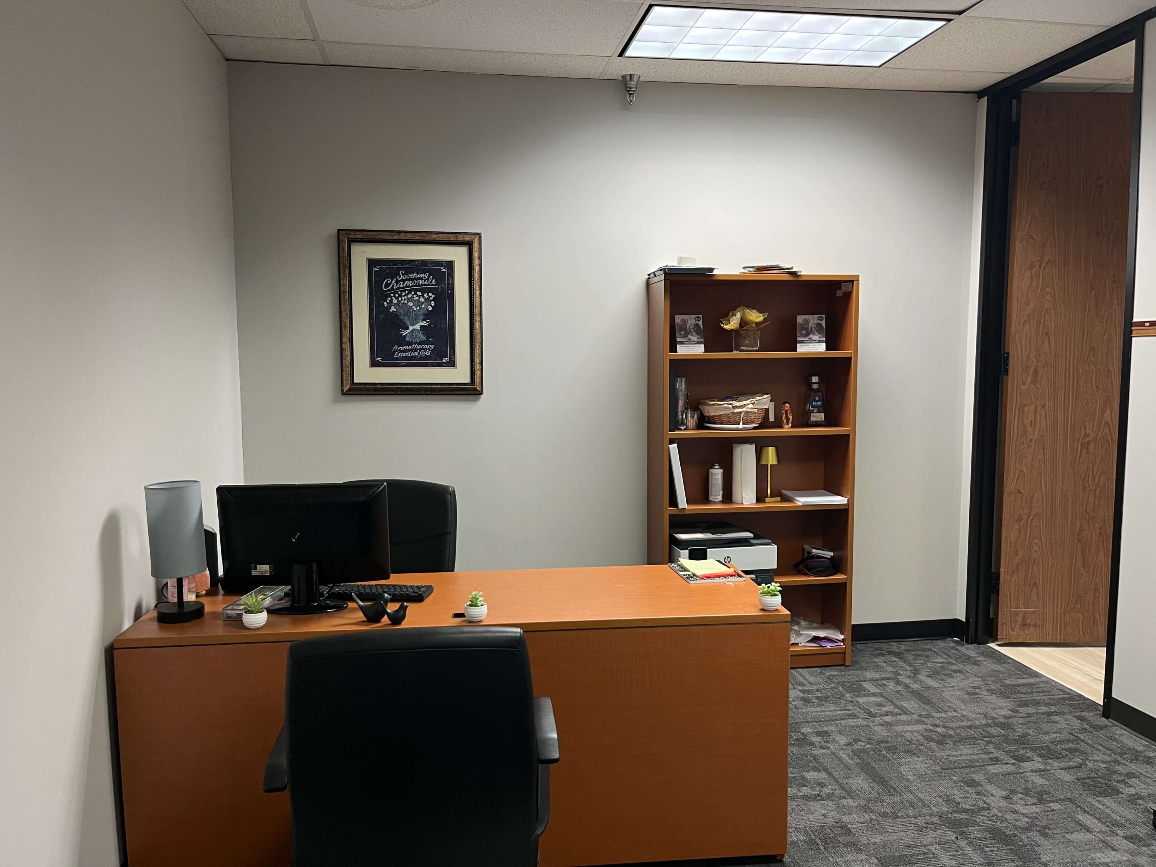 The image shows a small office space featuring a wooden desk with a computer and black chair, along with a bookshelf containing various items.