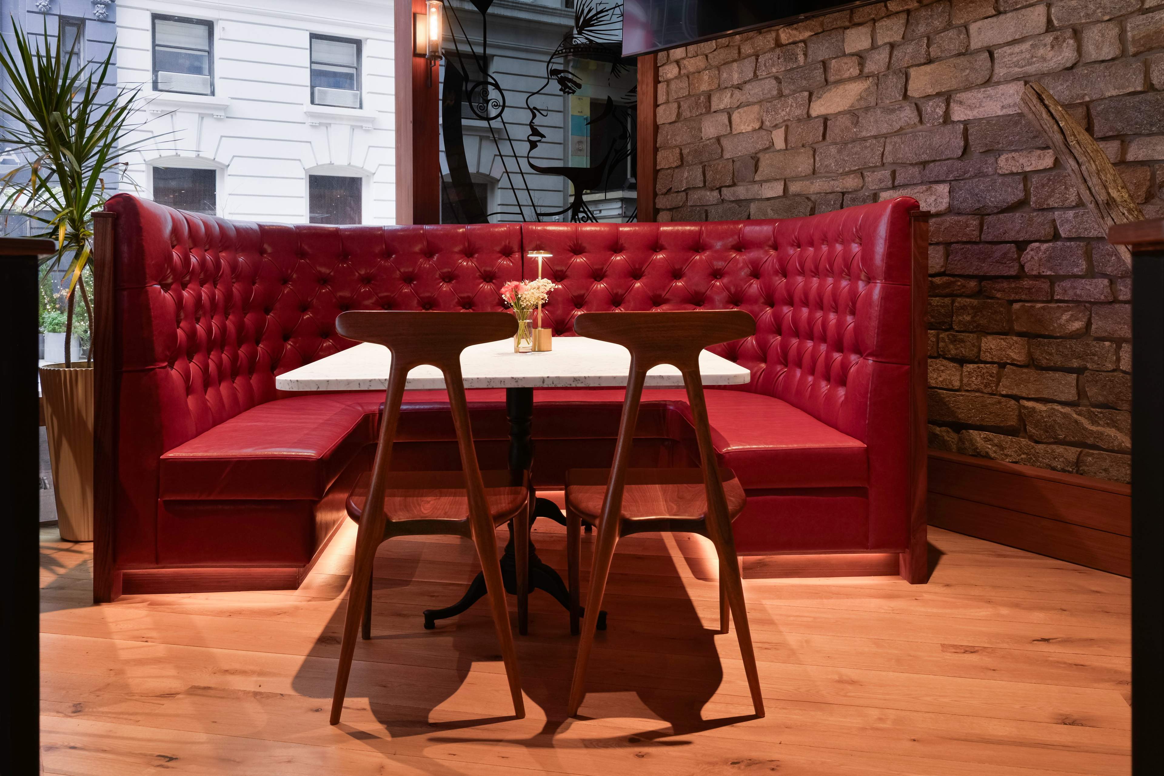 The image shows a cozy red upholstered booth with a small white table set for two, surrounded by wooden flooring and a textured stone wall.