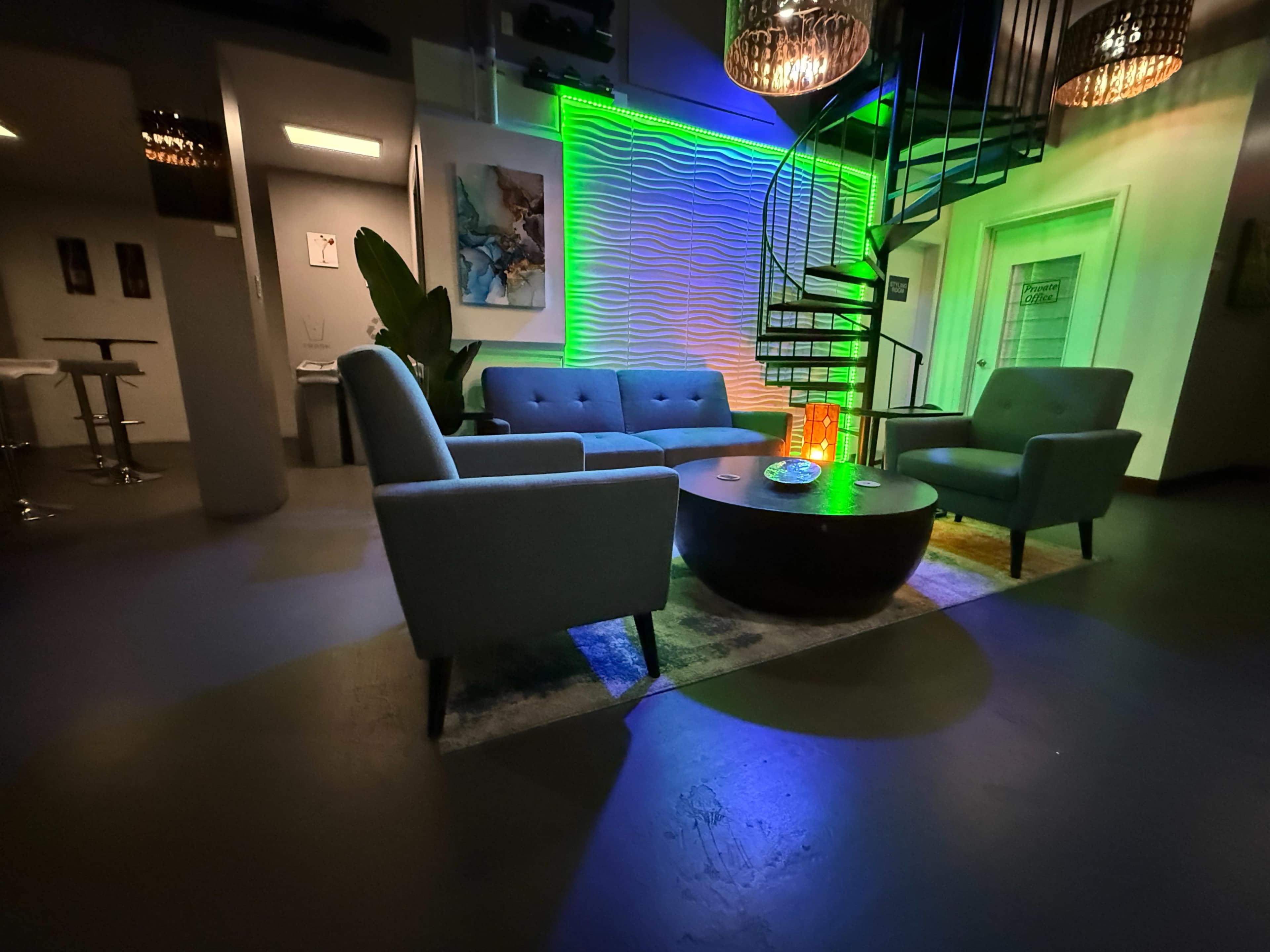 The image shows a modern lounge area featuring a spiral staircase, a round coffee table, and four upholstered chairs, with ambient green lighting on a textured wall.