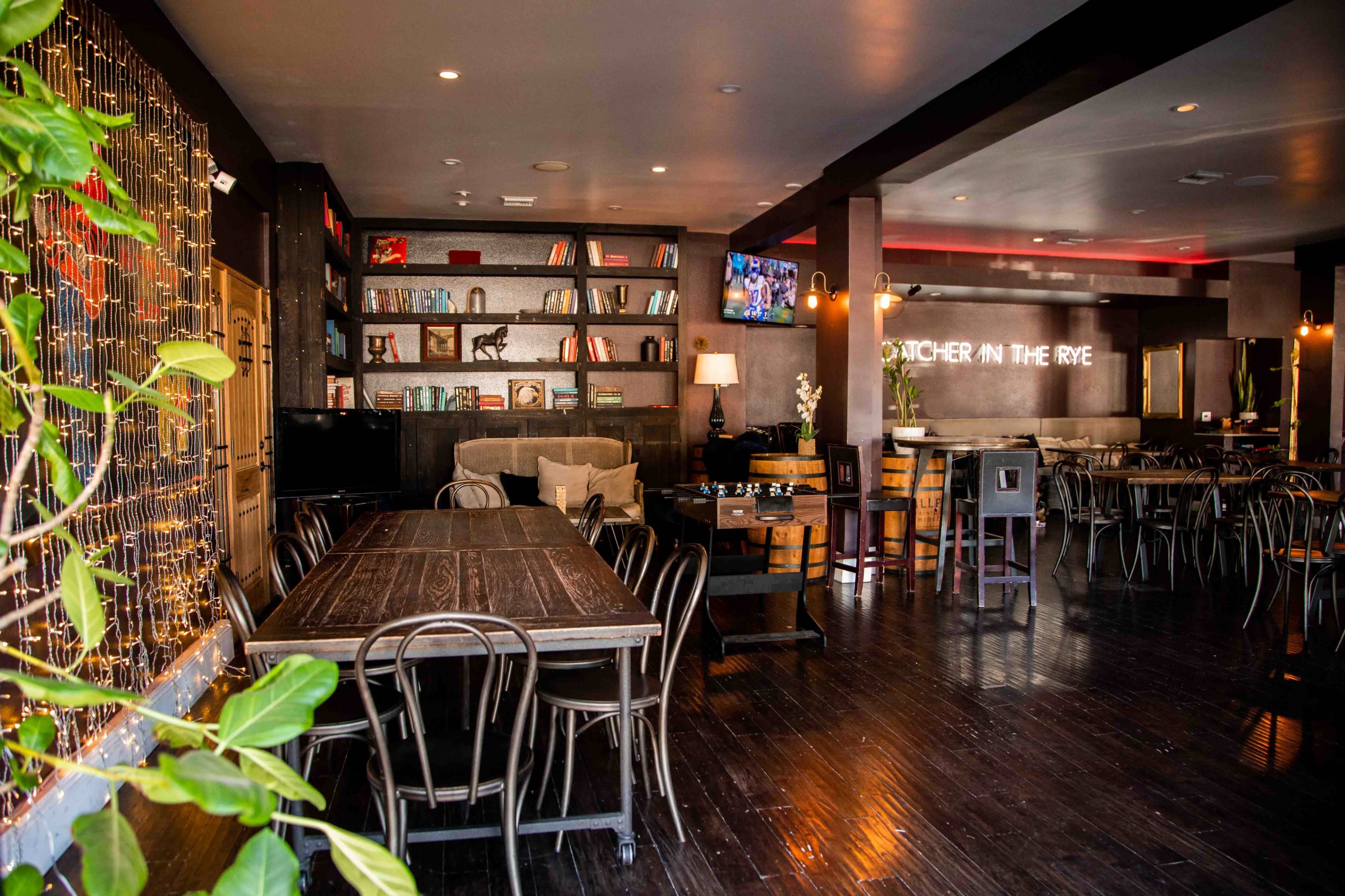 The interior of a modern restaurant features dark wooden tables, bookshelves filled with books, and a bar area illuminated by soft lighting.