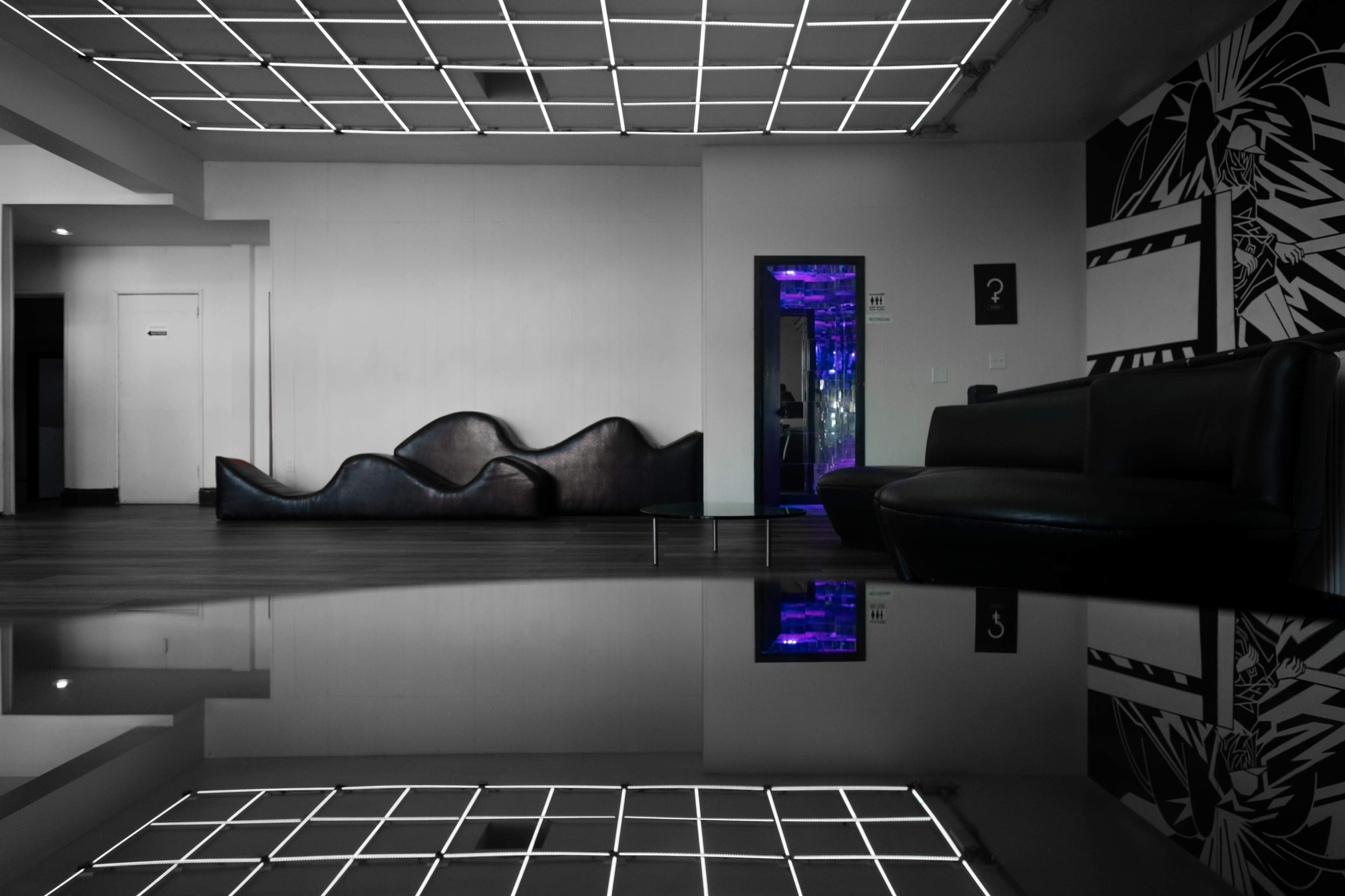 A spacious room with a wavy black sofa, a reflective floor, and a doorway leading to a colorful lighted area.