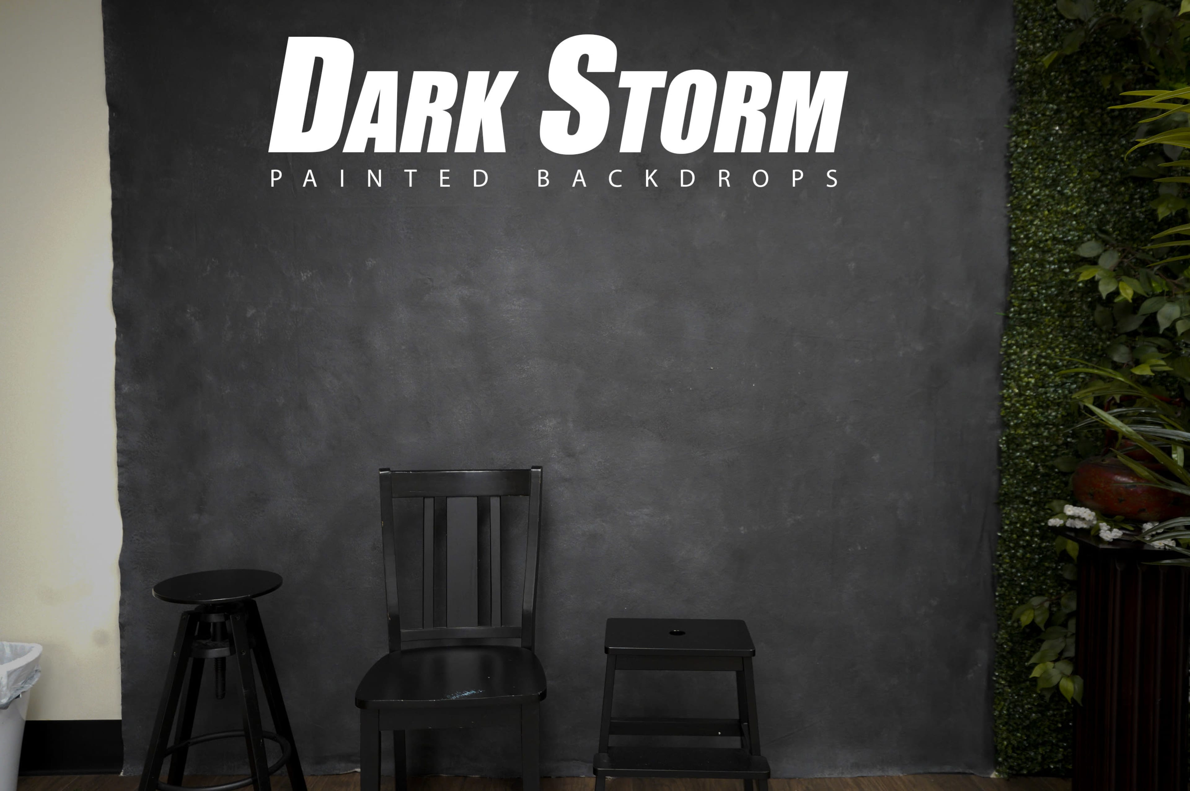 A minimalist setup featuring three black chairs positioned against a dark, textured backdrop with greenery on one side.