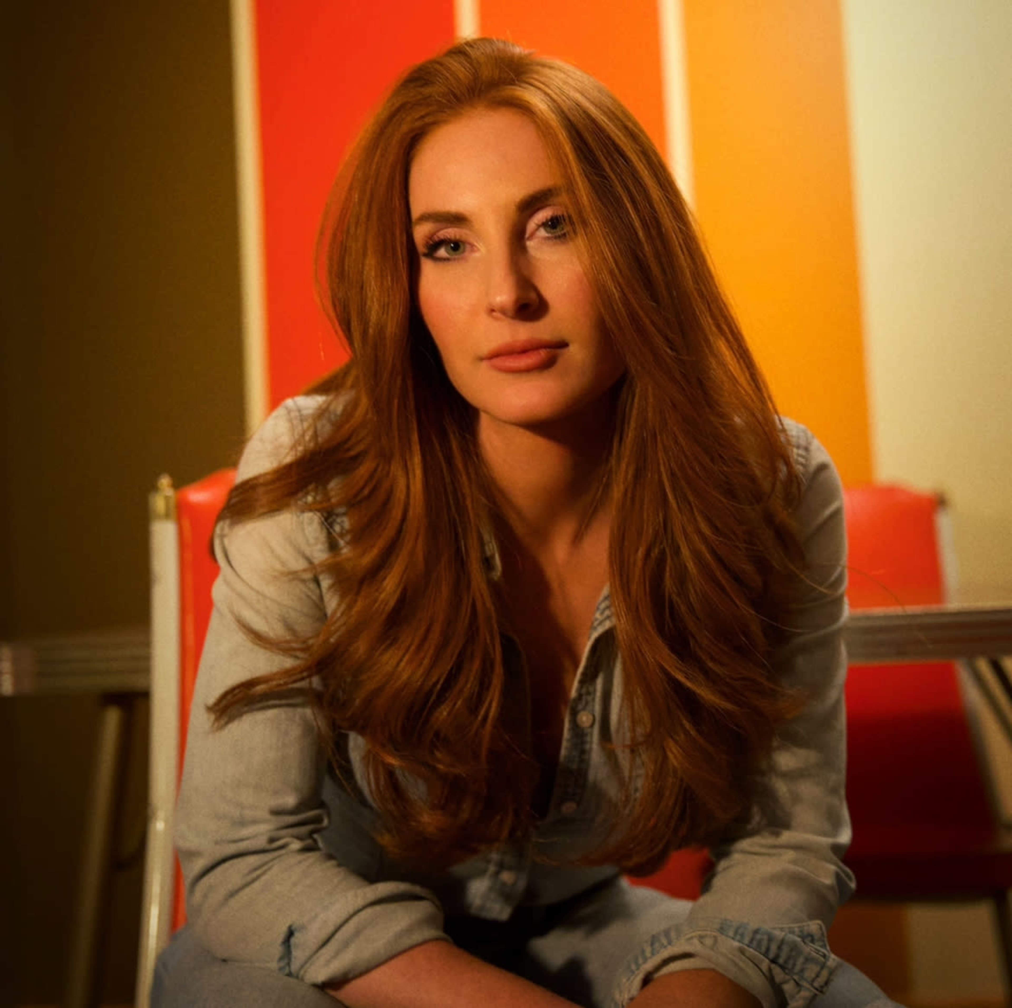 A woman with long, red hair sits on a chair in front of colorful backdrop panels.