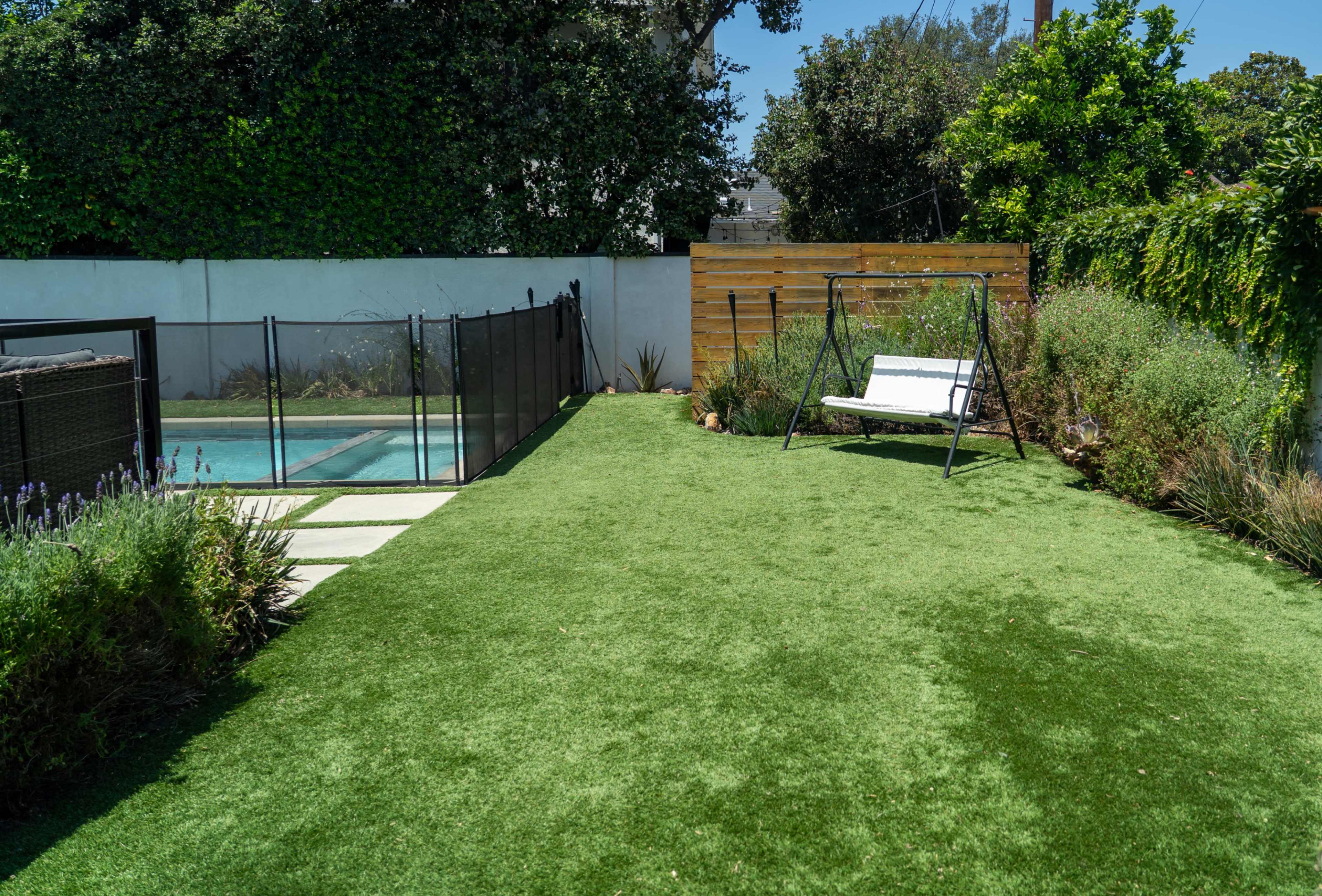 The image shows a well-maintained backyard featuring a pool, a grassy area, a wooden fence, and a swing set.