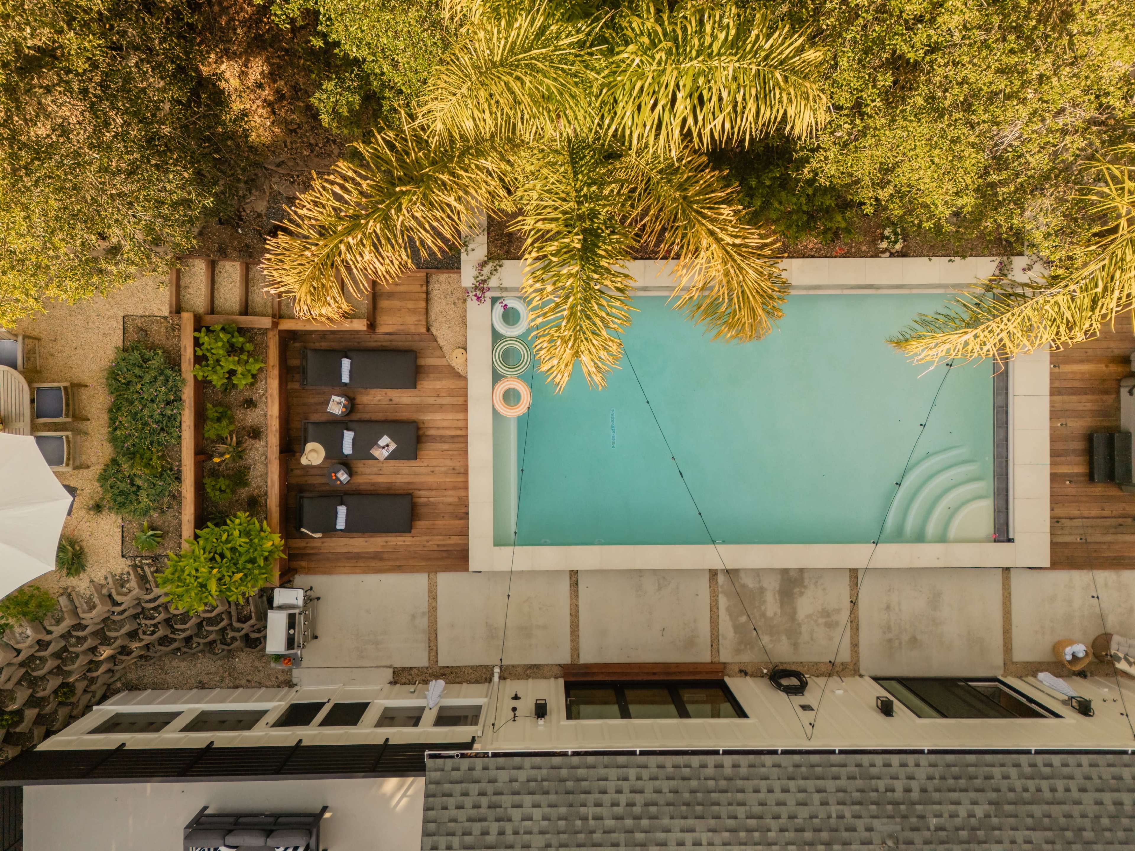 The image shows an aerial view of a backyard with a rectangular swimming pool surrounded by wooden decking, lounge chairs, and greenery.