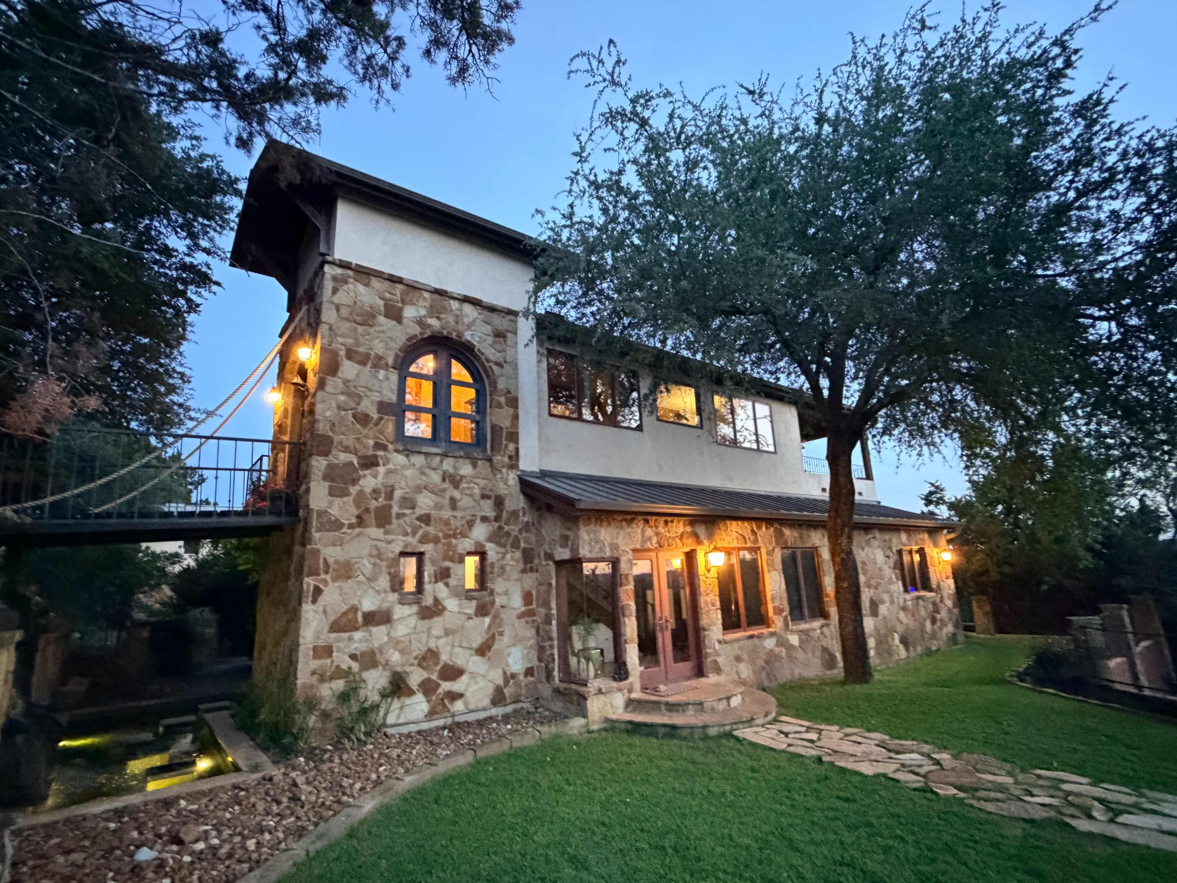 A stone house with a second-floor balcony is illuminated by warm lights and surrounded by green grass and trees at dusk.
