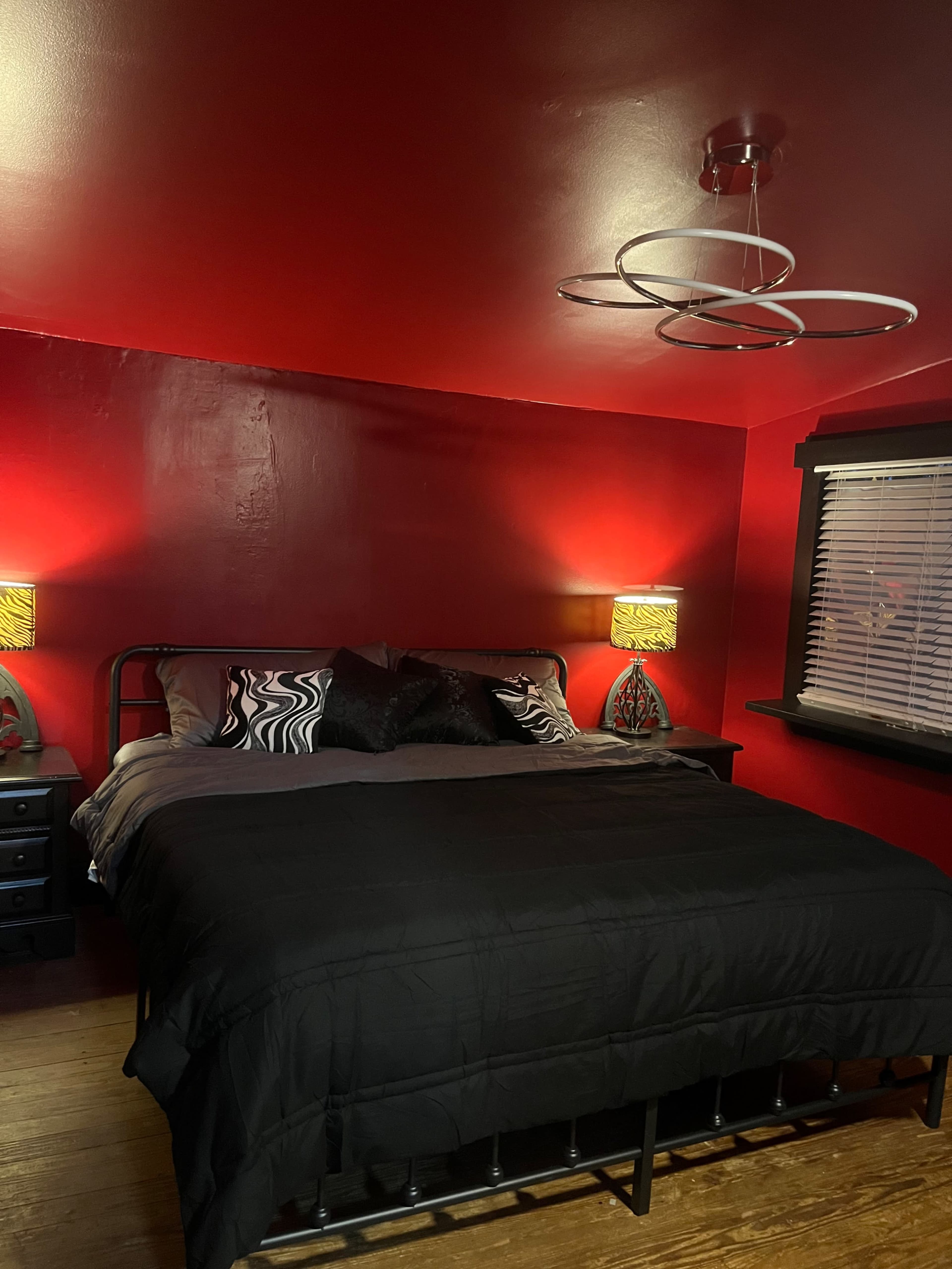 The image shows a small bedroom with a black metal bed dressed in dark bedding, flanked by decorative lamps on nightstands against red walls.