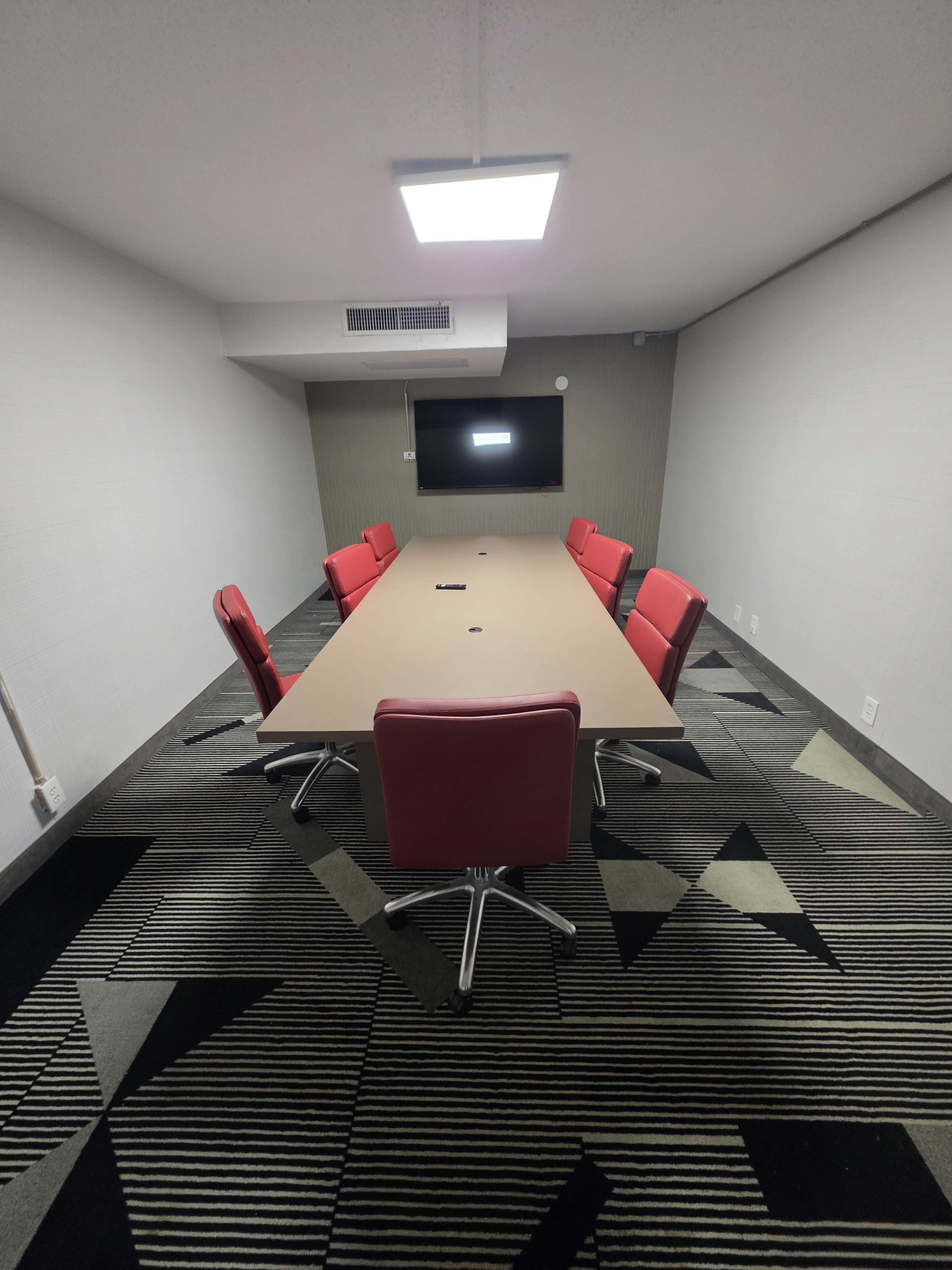 A rectangular conference table surrounded by six red chairs is set in a sparsely decorated meeting room with a dark patterned carpet and a wall-mounted screen.