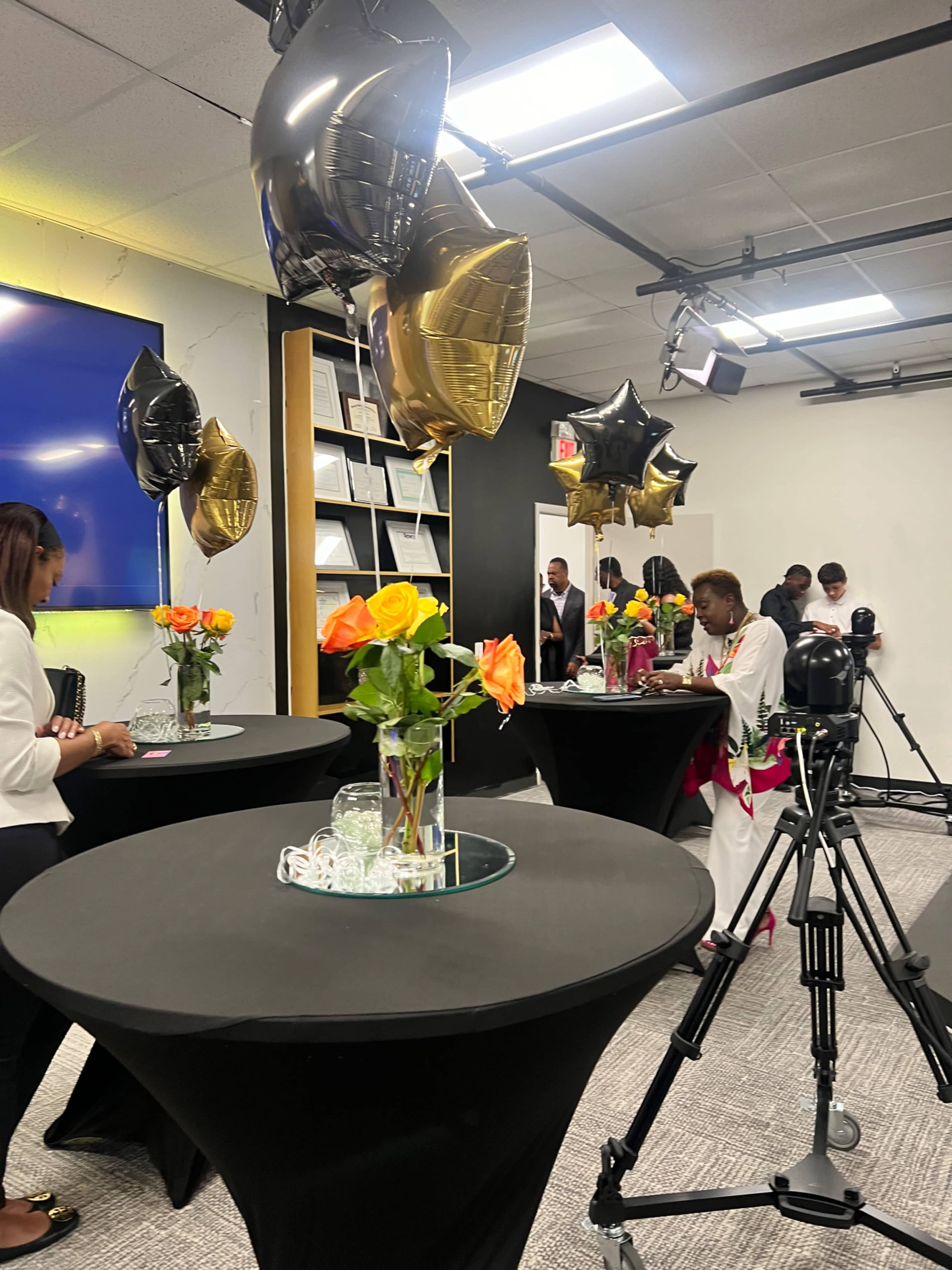 The image shows a decorated event space with black tablecloths, floral centerpieces, and balloons, where people are engaged in various activities.