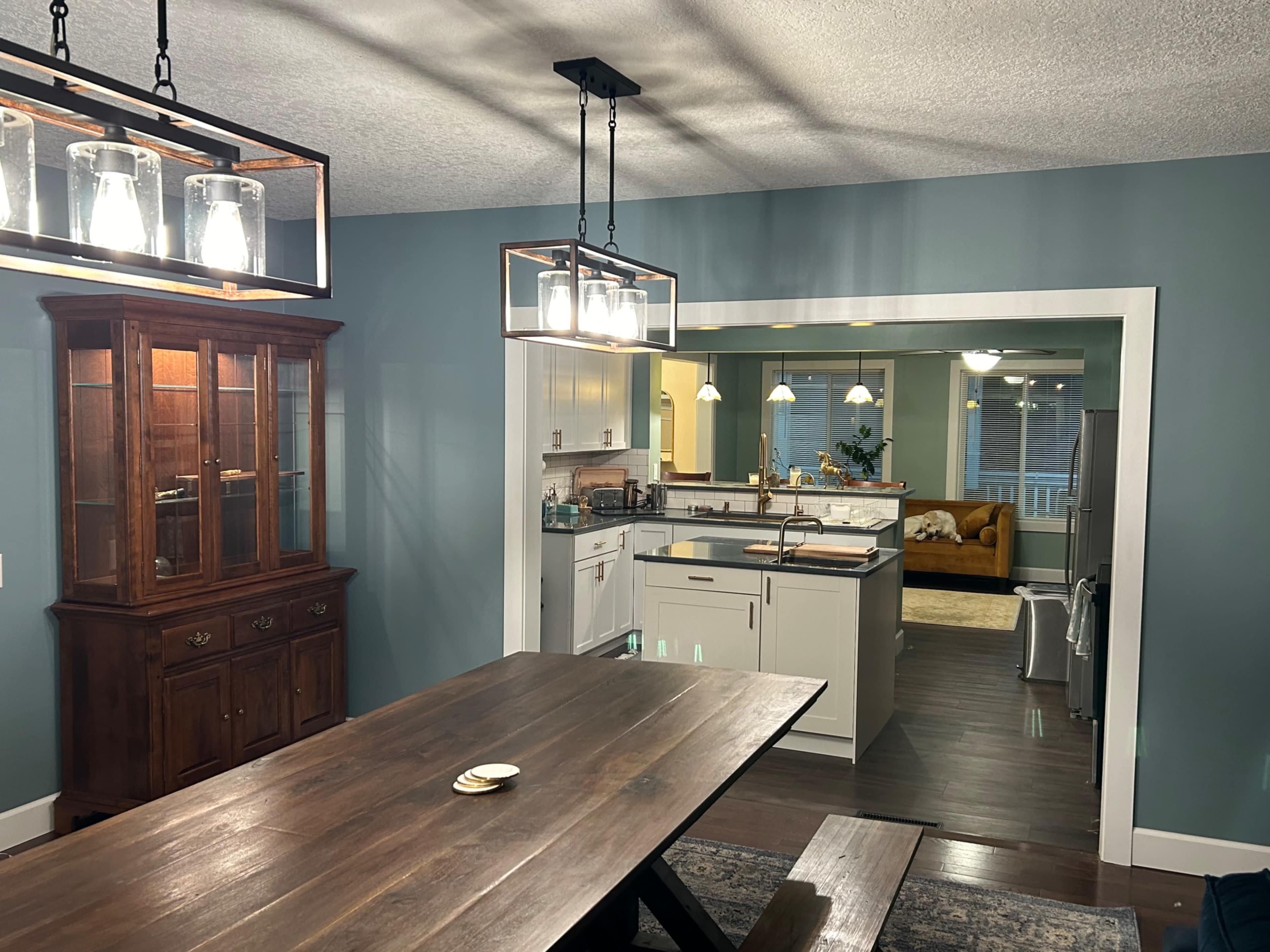 The image shows a dining area with a wooden table and a dark wood china cabinet, leading to a kitchen with white cabinets and visible appliances.