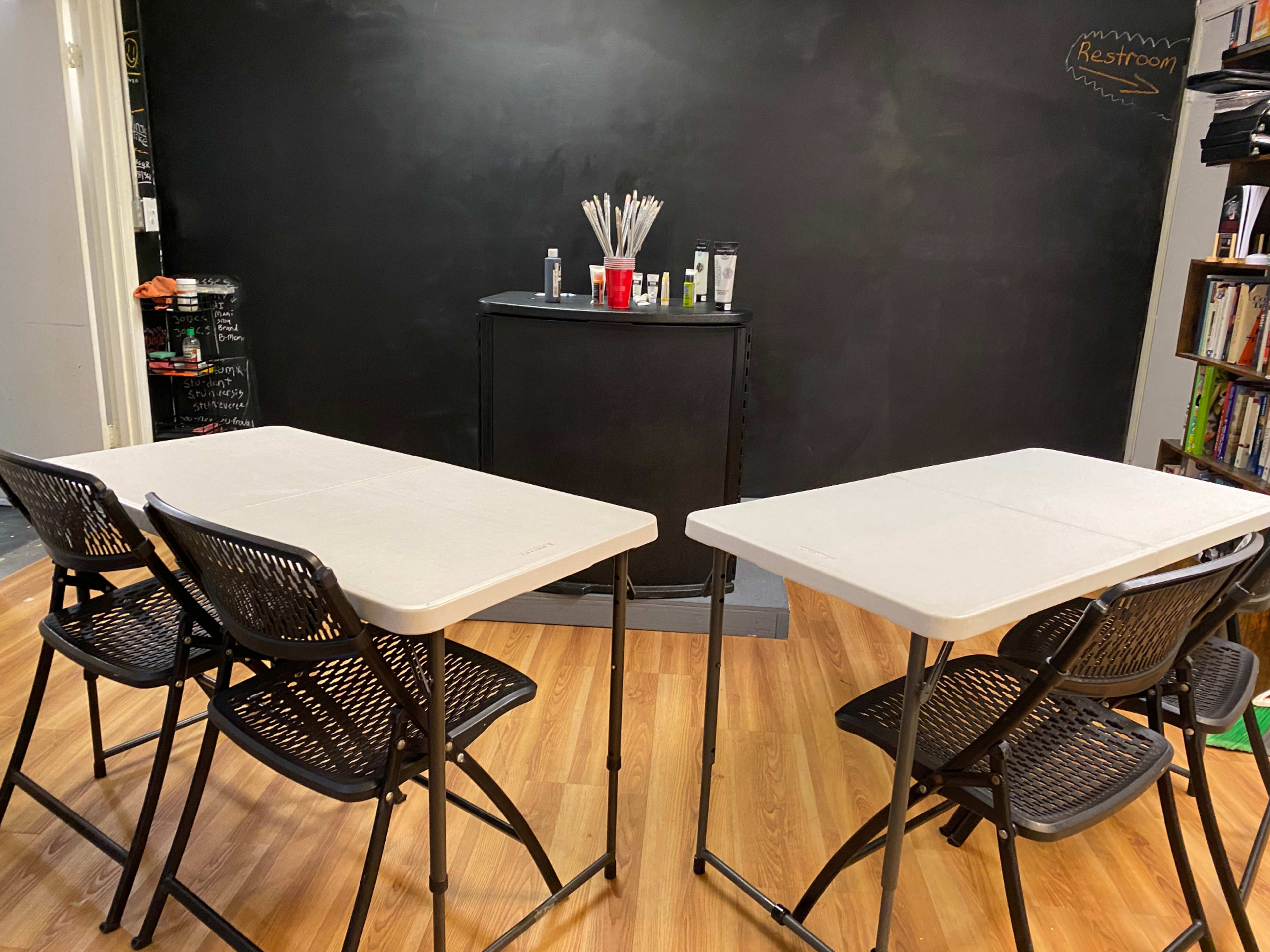 A classroom setup features two white tables with black folding chairs facing a blackboard and a counter with art supplies.