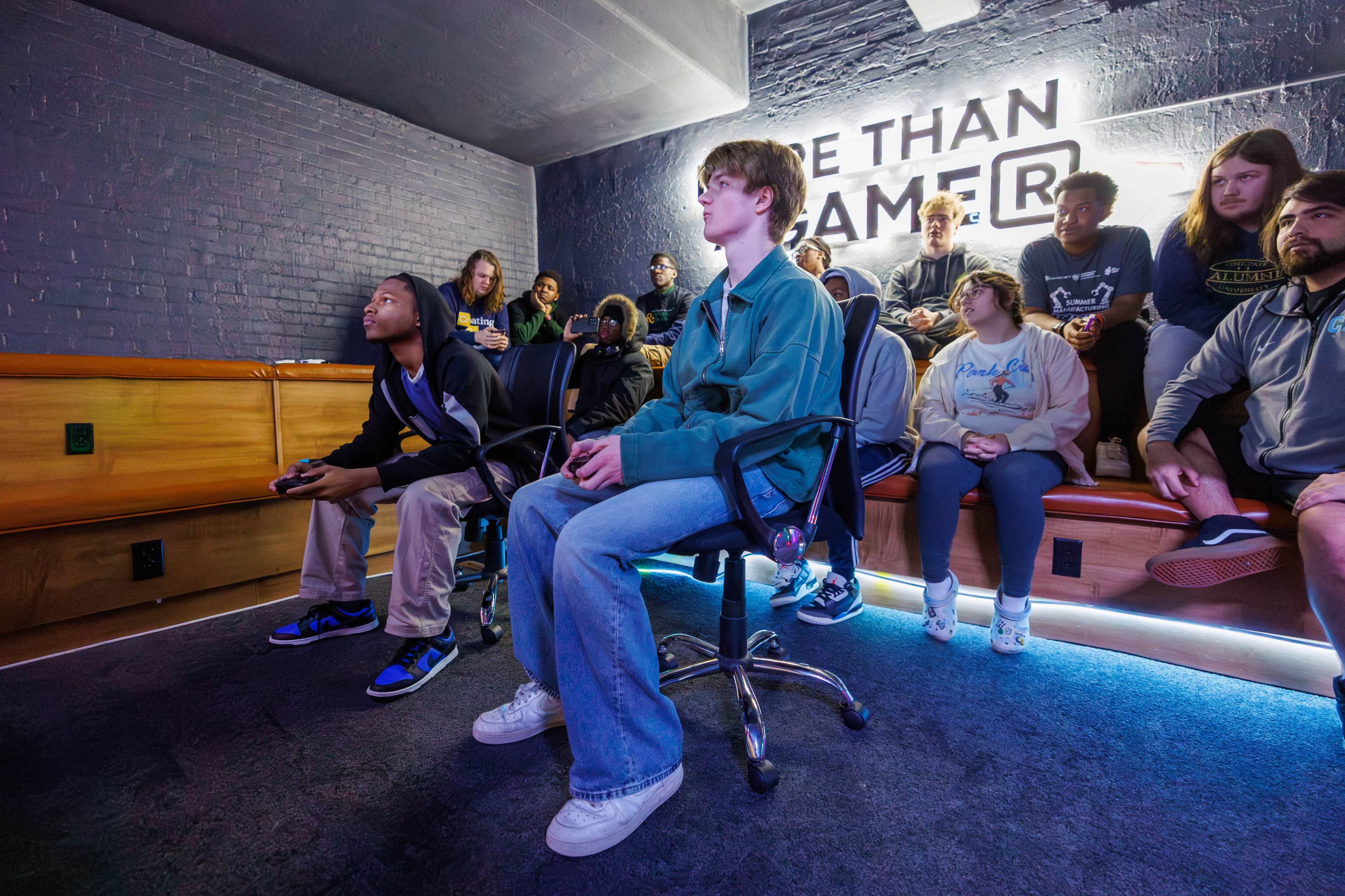 A group of young adults watches intently as two players compete in a video game in a dimly lit room.