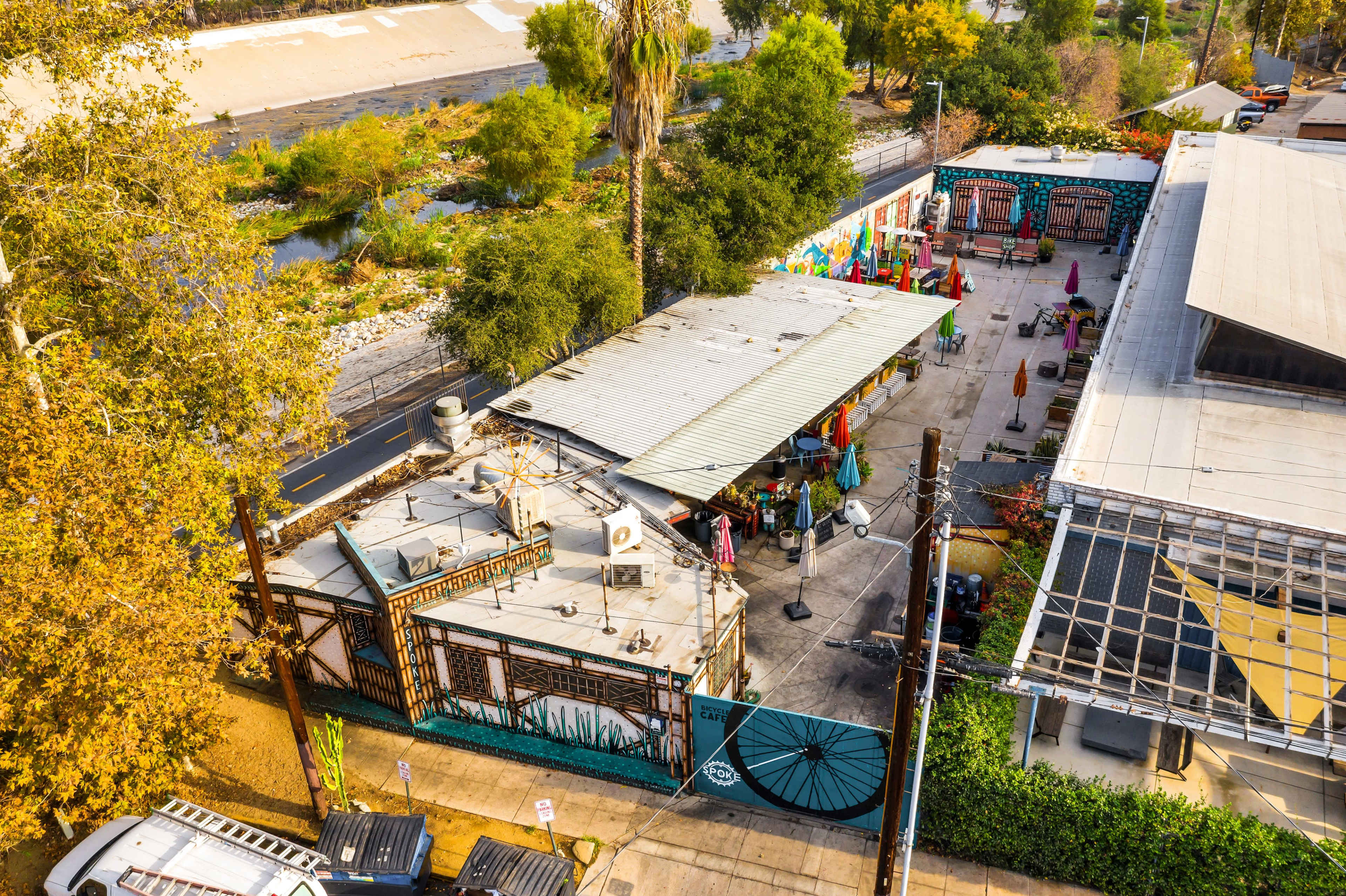 A colorful outdoor market area with various tents and vendors, surrounded by lush trees and urban buildings.